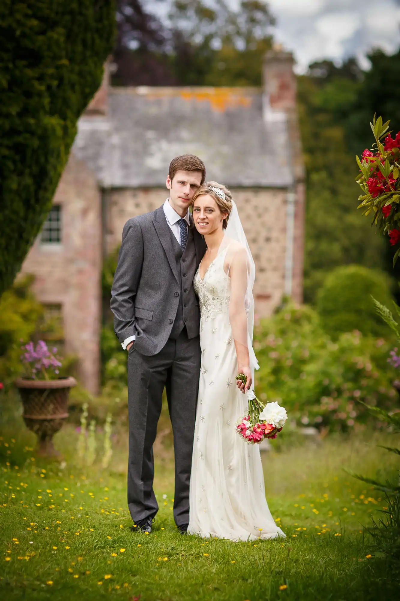 A bride and groom stand together in a garden, with the bride holding a bouquet and the groom in a gray suit, with a historic stone building in the background.