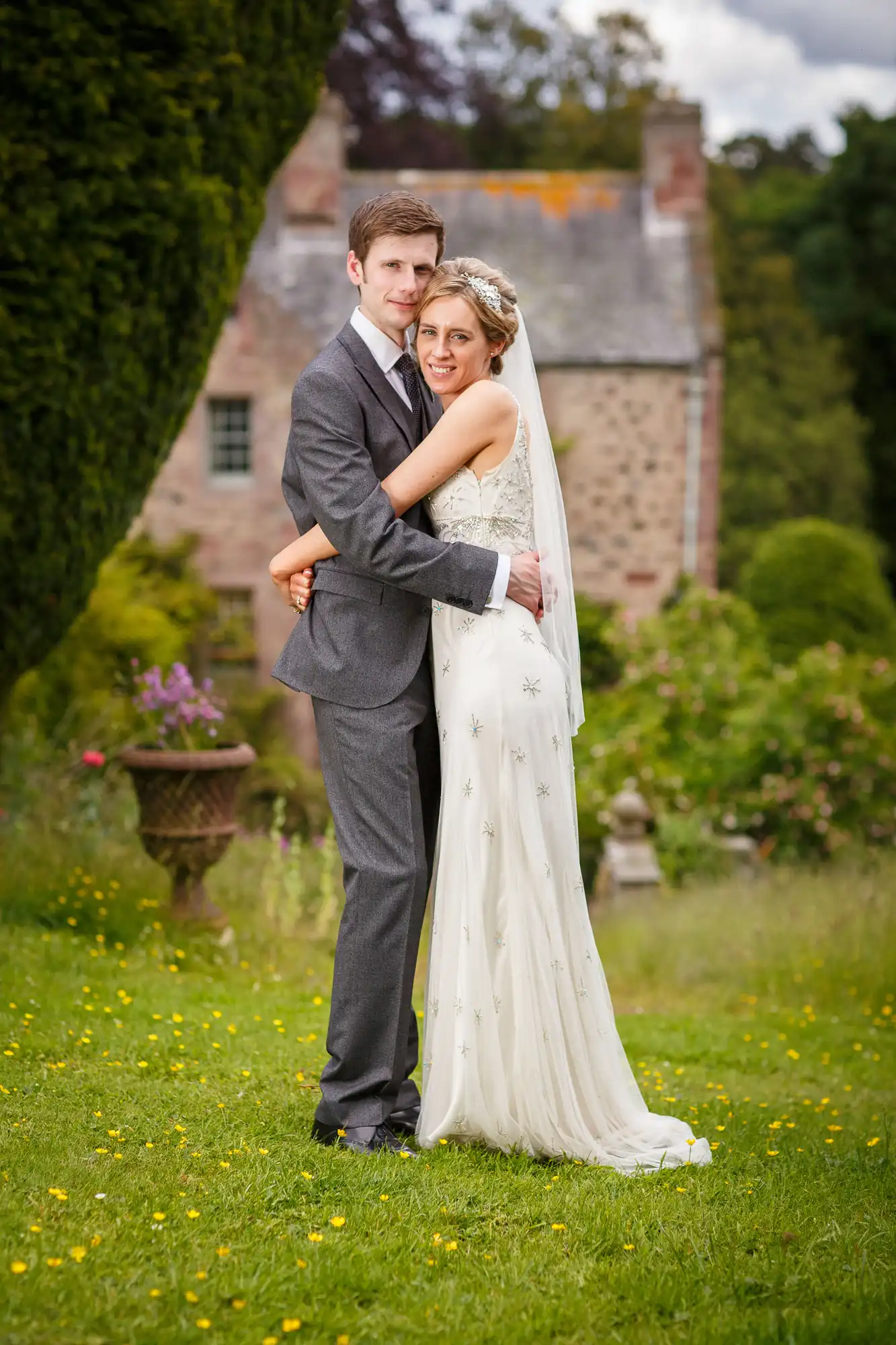 A bride in a white gown and a groom in a gray suit embrace in a garden with a historic brick building in the background.
