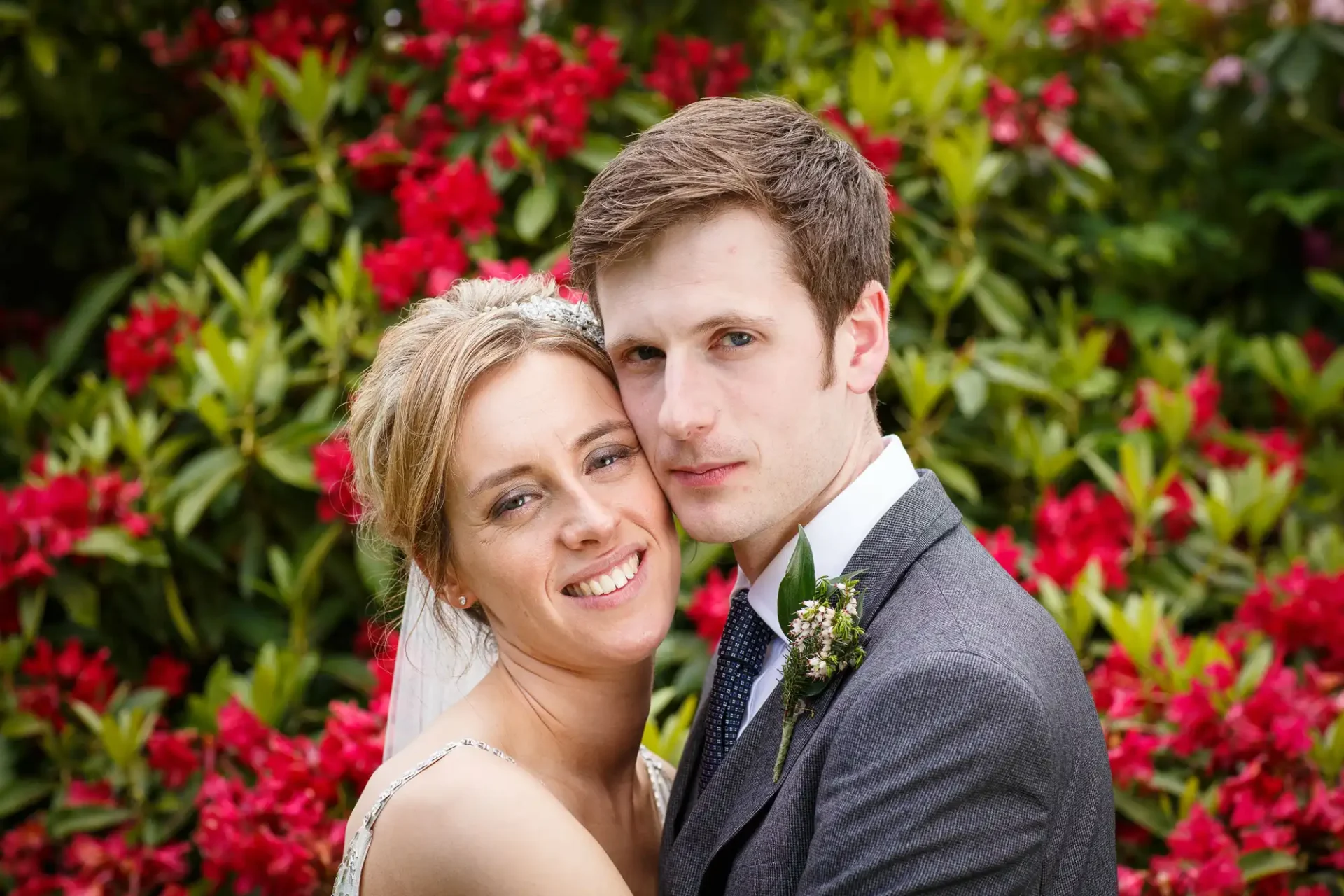 A bride and groom posing affectionately in front of vibrant red flowers.