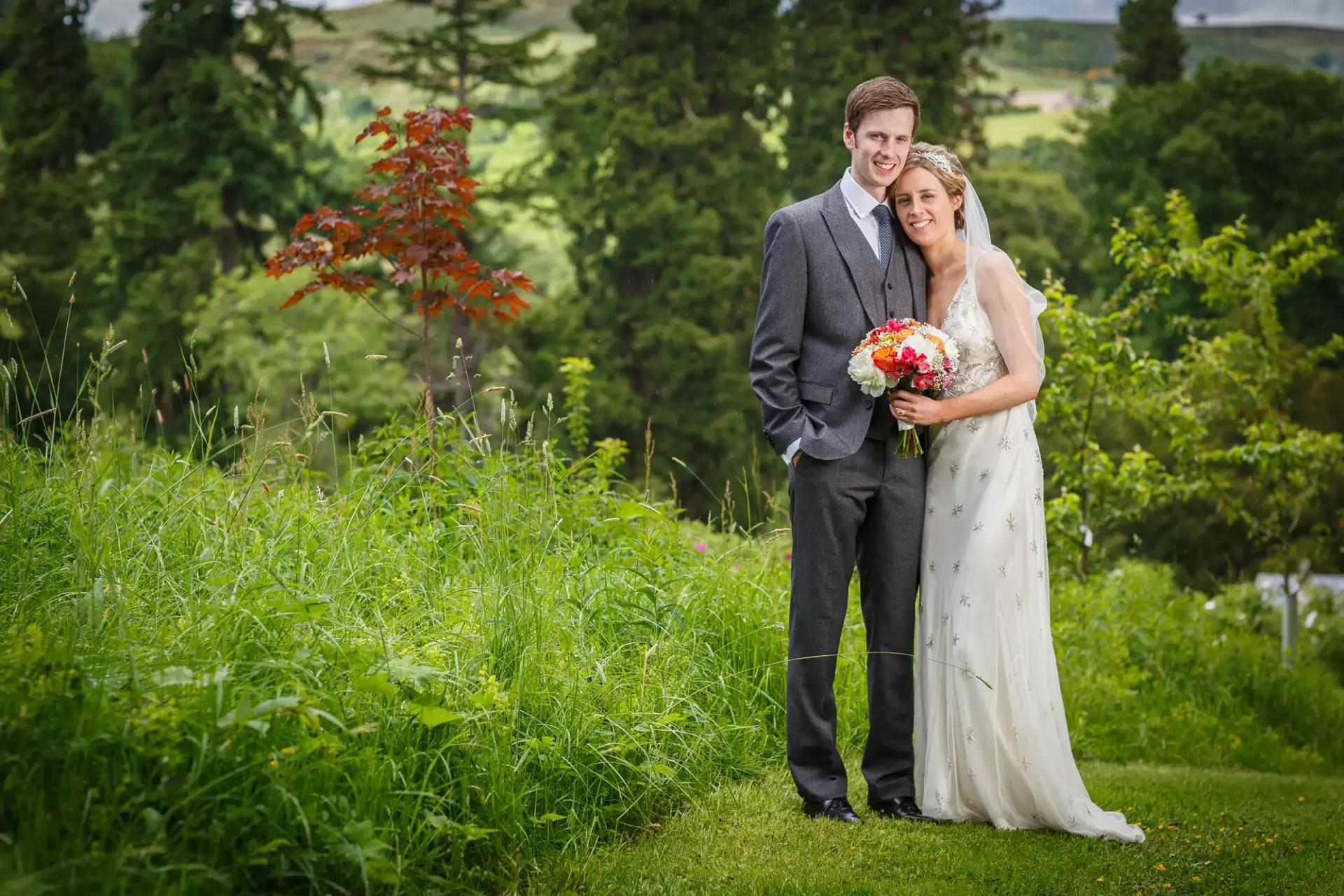 A bride and groom smiling, standing in a lush green field with trees in the background, the bride holding a bouquet.