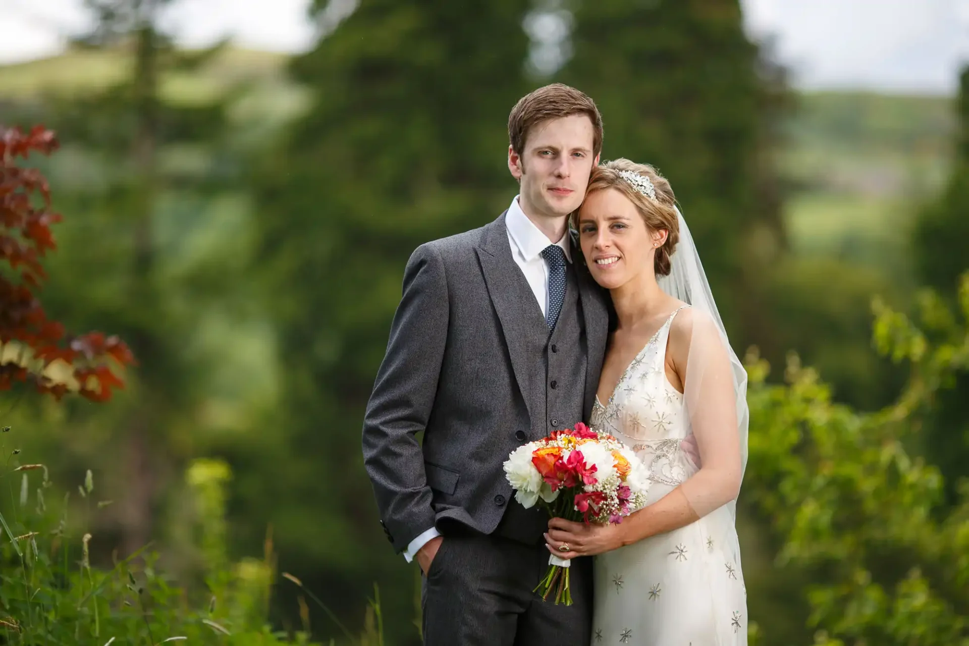 A bride and groom posing outdoors with a bouquet, set against a lush green background with trees.