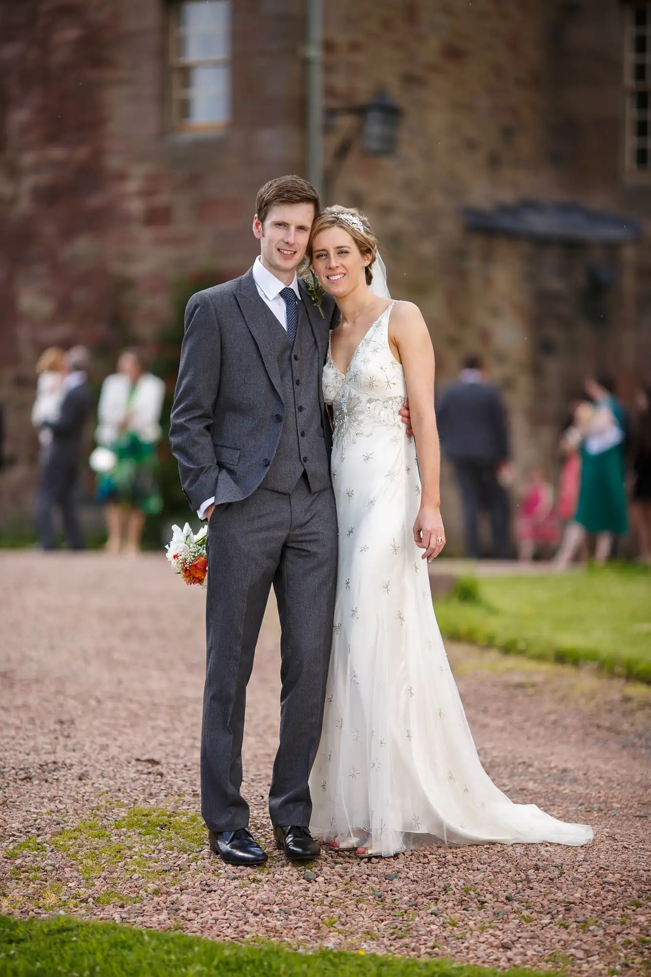 A bride and groom smiling outdoors, the bride in a white dress and the groom in a gray suit, standing on a gravel path with a castle and guests in the background.
