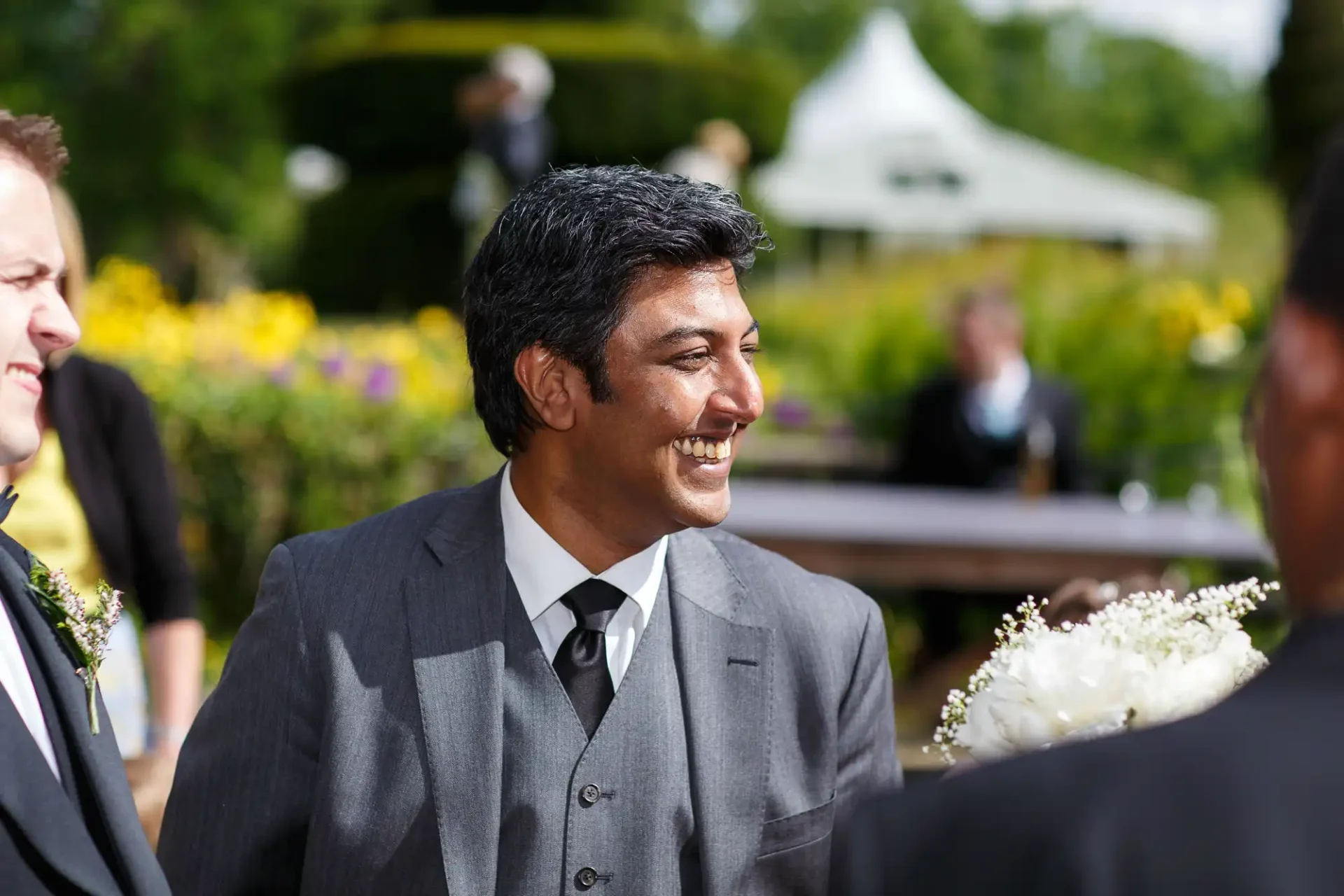 A man in a gray suit smiling at a social gathering outdoors, surrounded by lush greenery and bright flowers.