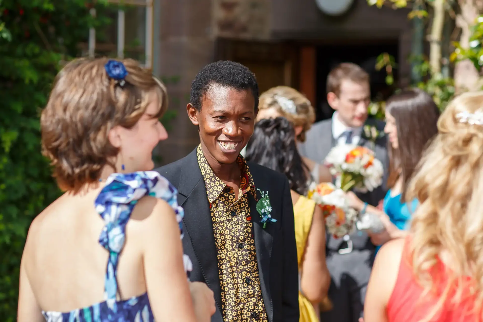 A smiling person in a patterned gold and black outfit interacts with guests at a sunny outdoor wedding.