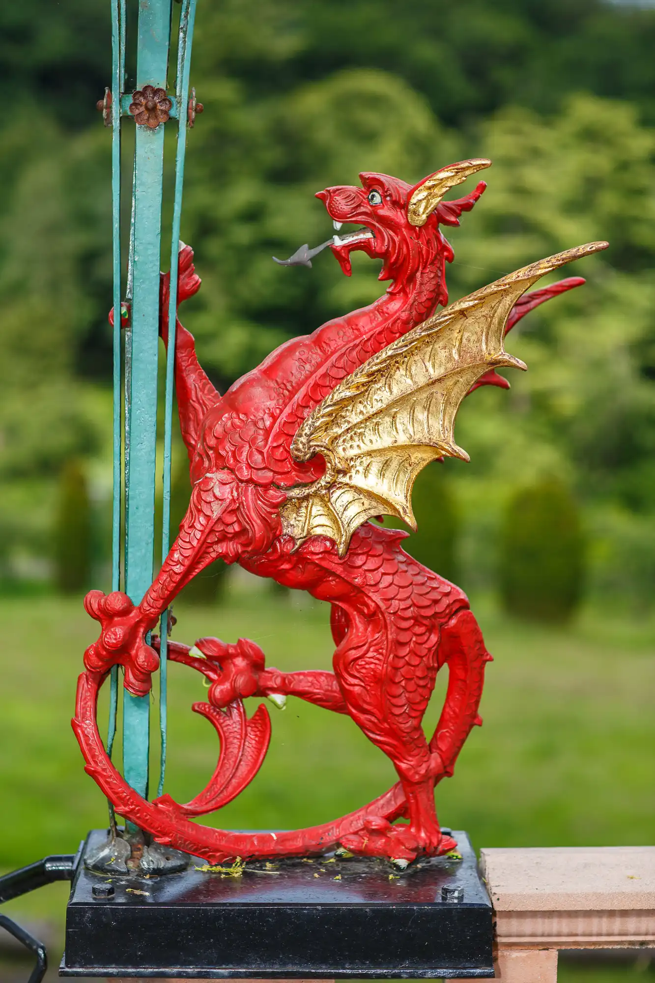 A vibrant red and gold dragon statue with wings spread and mouth open, mounted on a fence against a blurred green background.