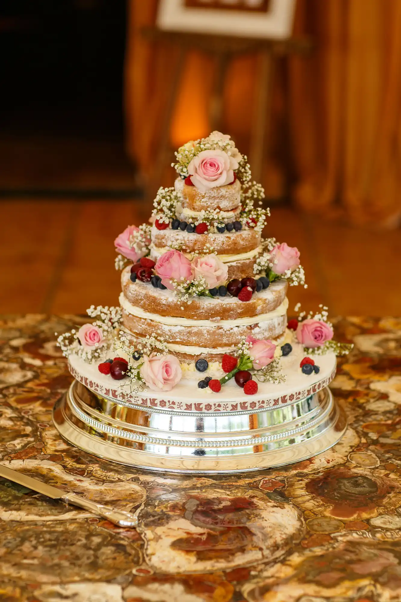 A multi-tiered naked wedding cake adorned with fresh berries, pink roses, and small white flowers, displayed on a decorative silver stand.