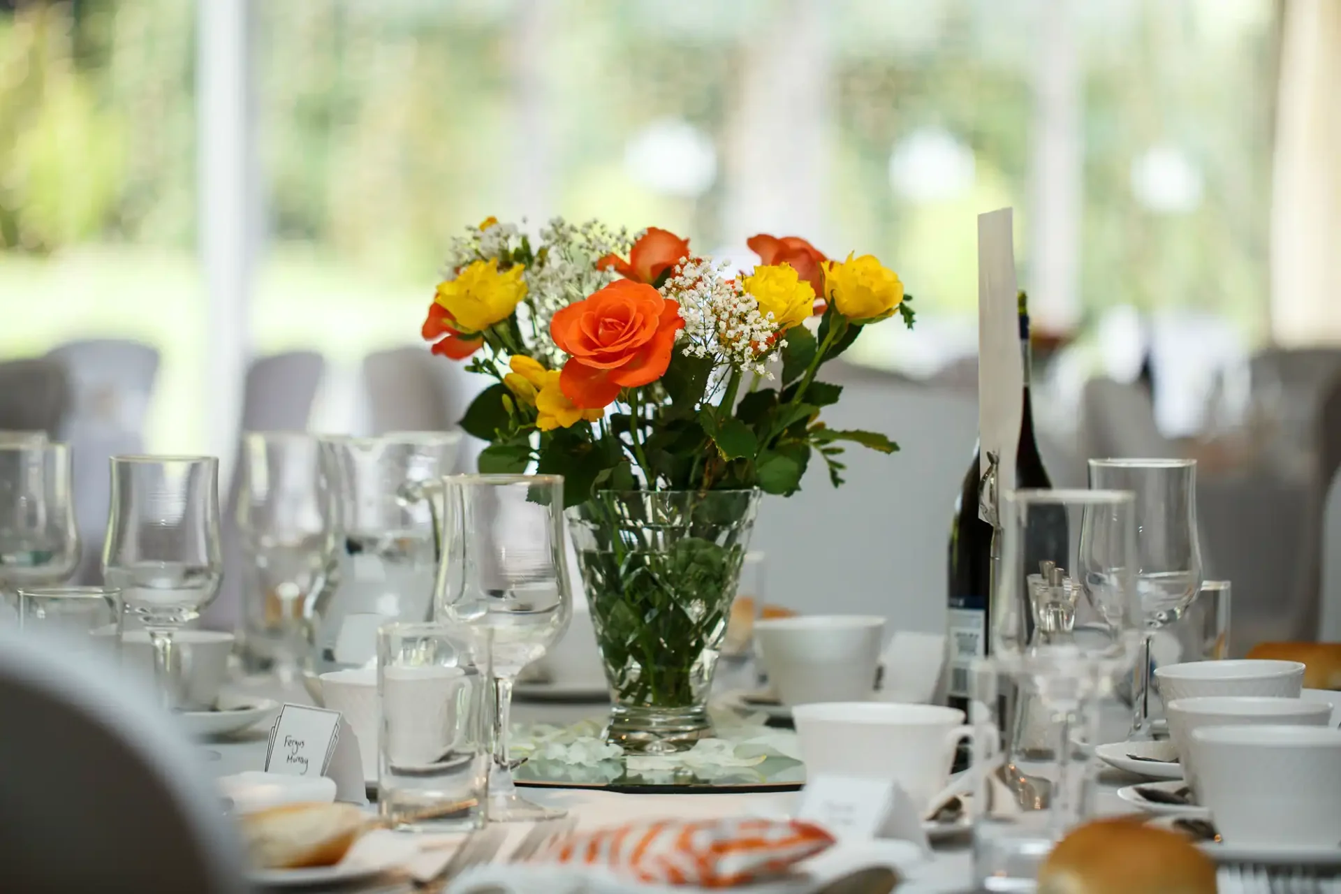 A table set for an event with a vase of yellow and orange flowers surrounded by glasses, plates, and cutlery.