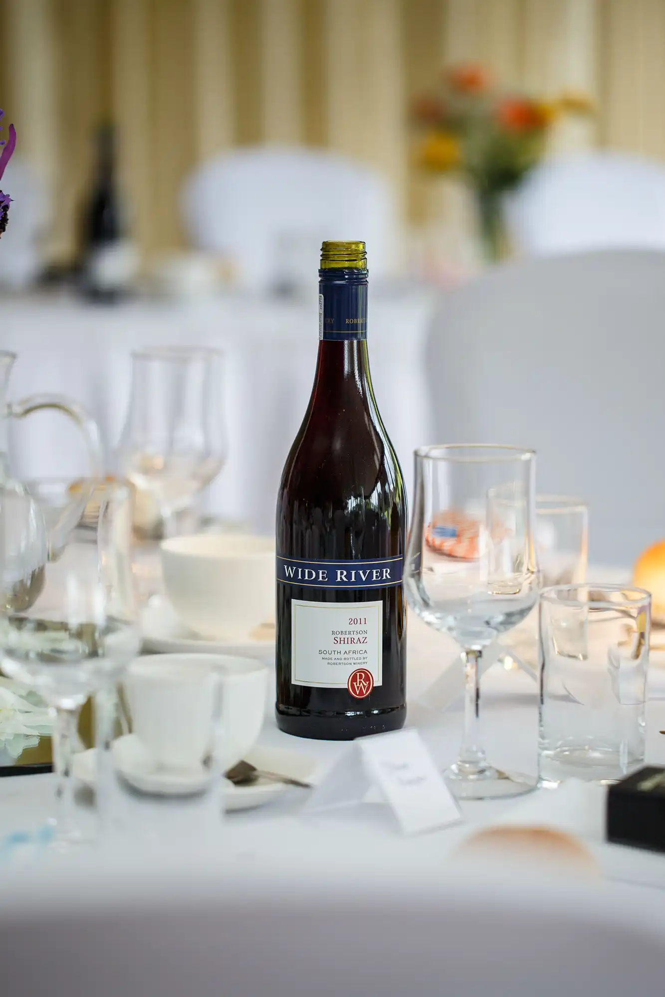 A bottle of "wide river" shiraz wine from 2017 stands on a dining table amid empty glasses and white chair covers.