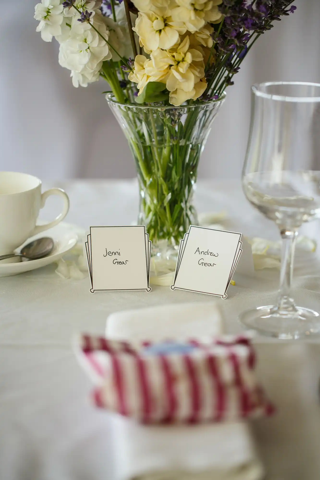Elegant dining table setup featuring a bouquet of flowers in a vase, name cards for guests, a white teacup, and a napkin wrapped with a pink-striped ribbon.