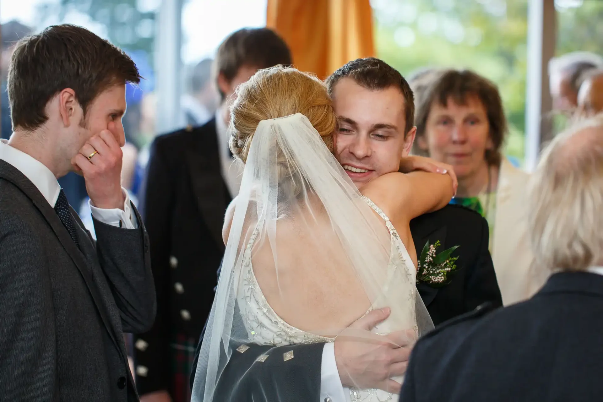 A groom hugs a bride wearing a veil at their wedding, surrounded by emotional guests.