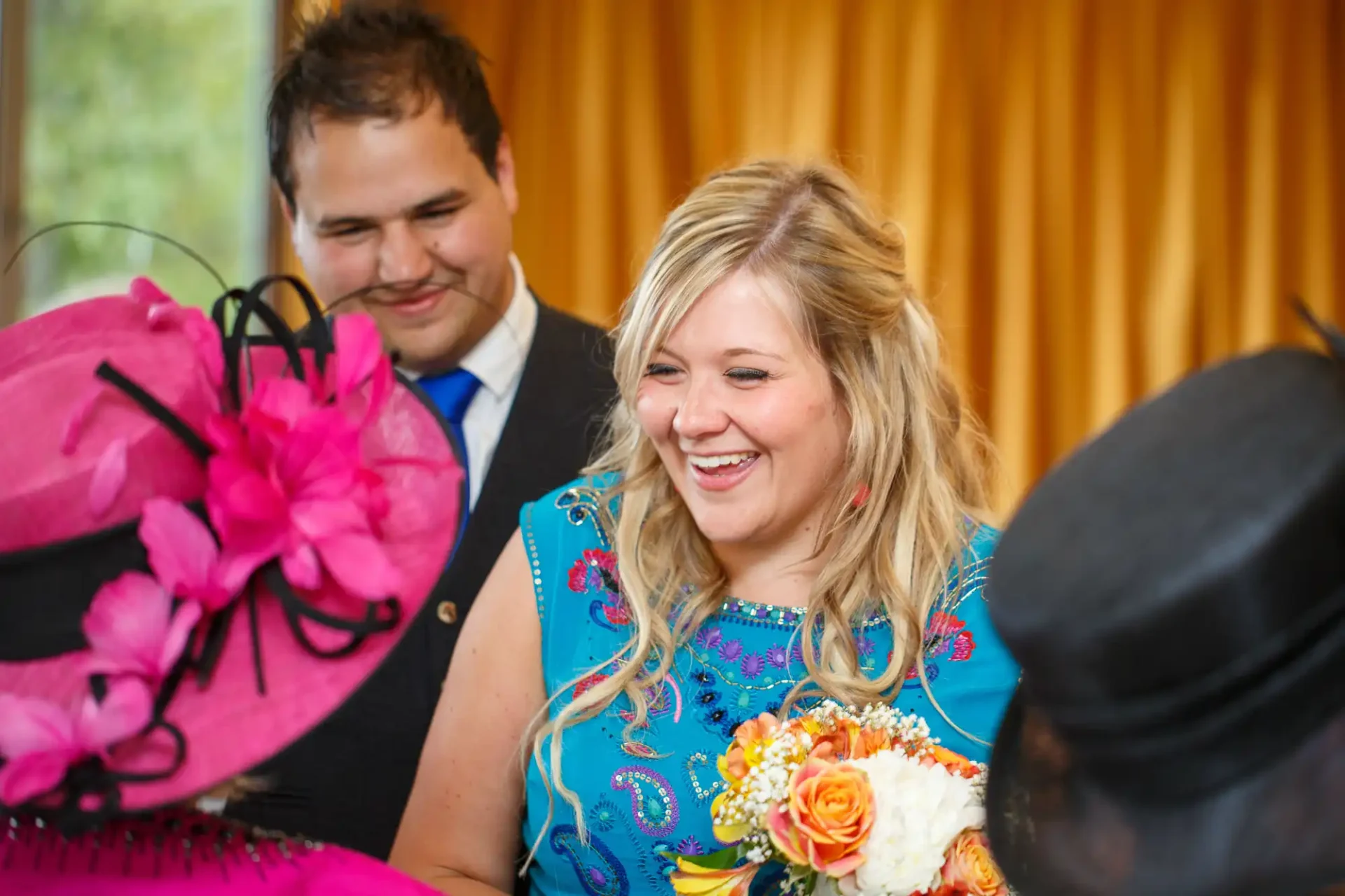 A woman in a blue dress laughs joyfully, holding flowers, with a man smiling in the background at a festive event.