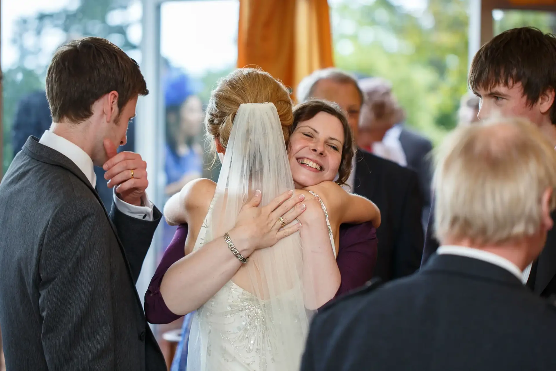 Bride embracing a woman in a blue dress at a wedding, surrounded by guests in a joyous atmosphere.
