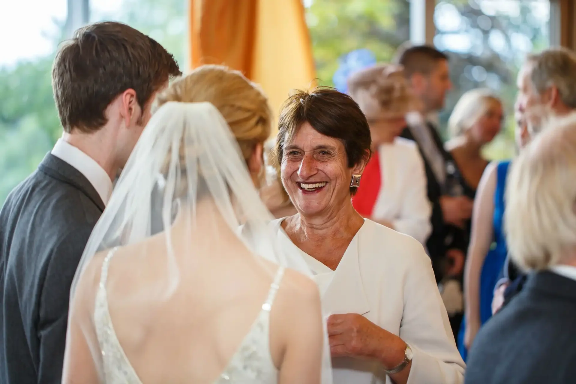 A joyful elderly woman chats with a bride and a groom at a wedding reception filled with guests in the background.