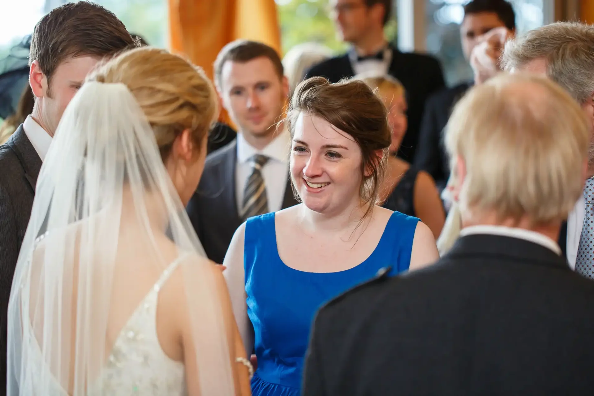 A woman in a blue dress smiling at a bride during a wedding ceremony, surrounded by guests.