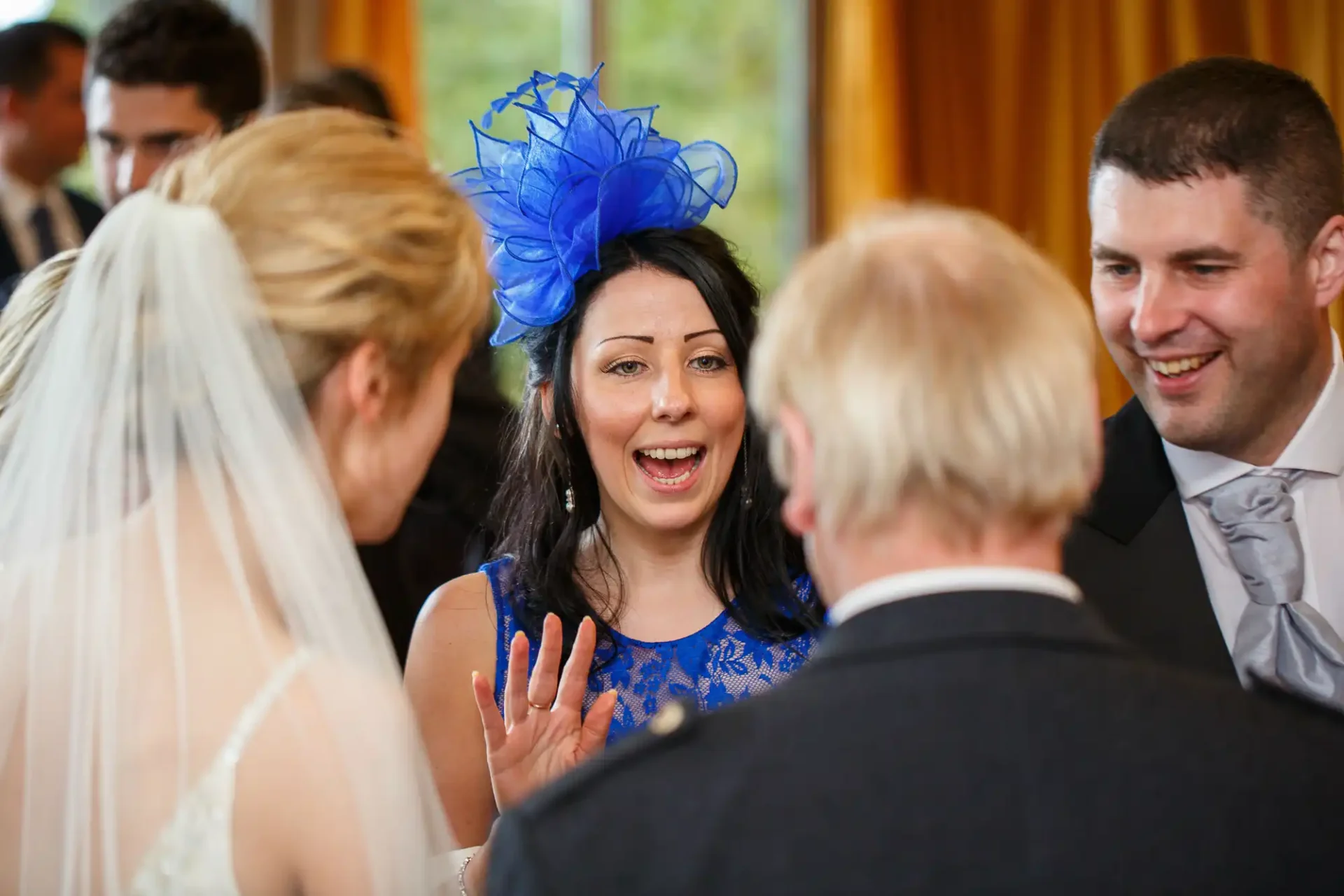 A woman in a blue dress and large blue hat laughing and talking with a bride and two men at a wedding reception.