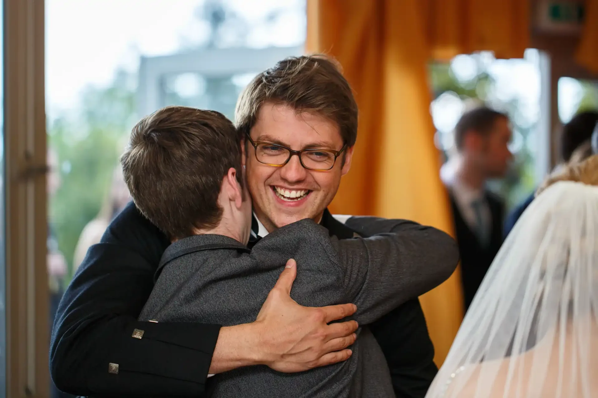 Two men embracing joyfully at a wedding reception, one facing the camera with a big smile.