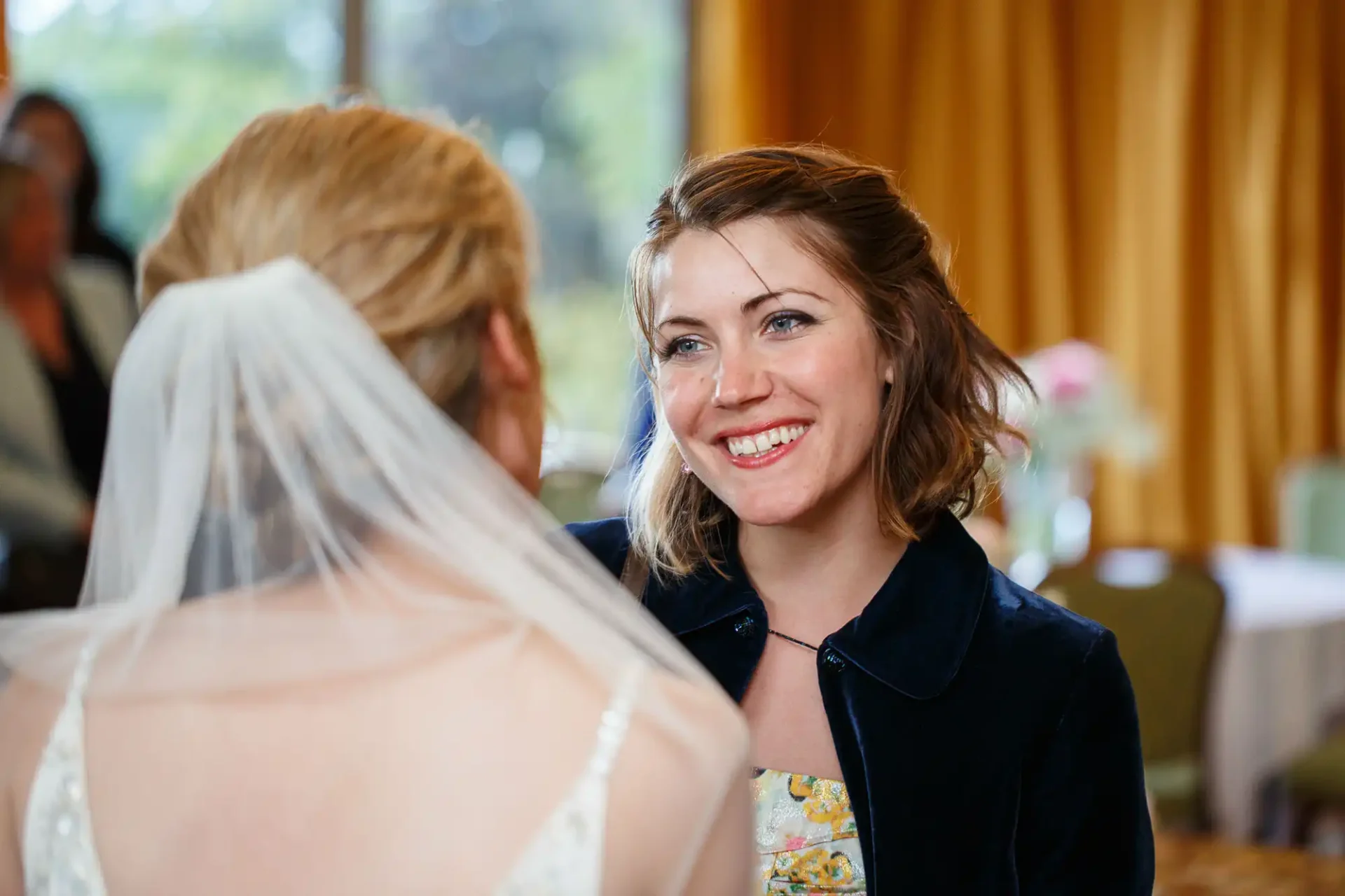A smiling woman in a floral dress and black jacket converses with a bride whose back is to the camera, in a room with elegant decor.