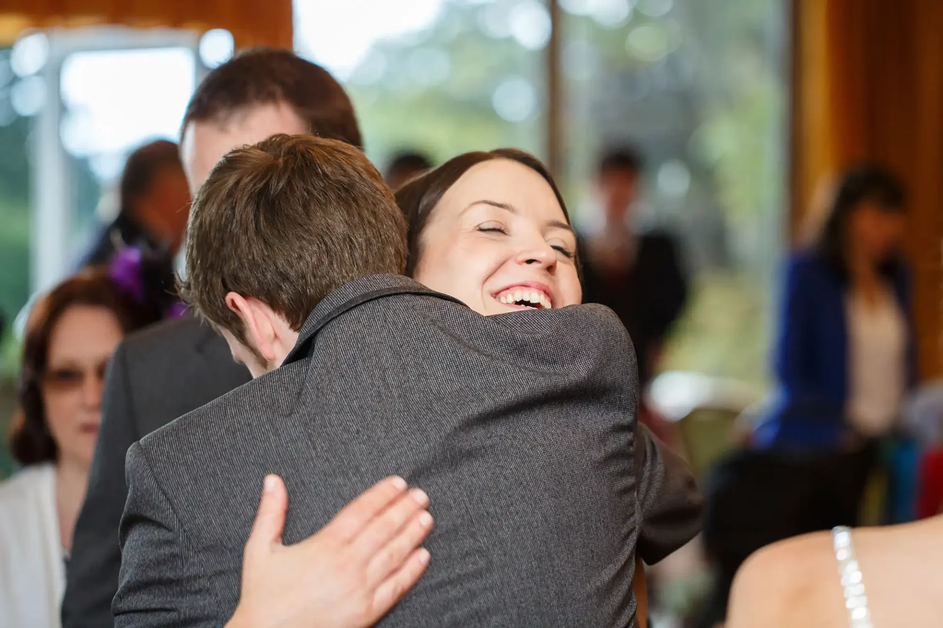 Woman smiling broadly while hugging a man at a social gathering, with other people in soft focus in the background.