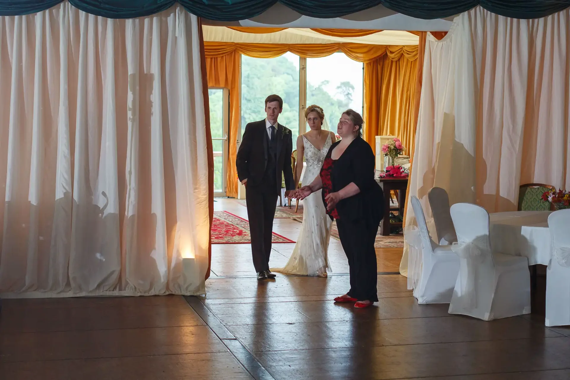 A bride and groom being escorted into a wedding reception by a woman in a black and red dress, with white chairs and curtains in the background.