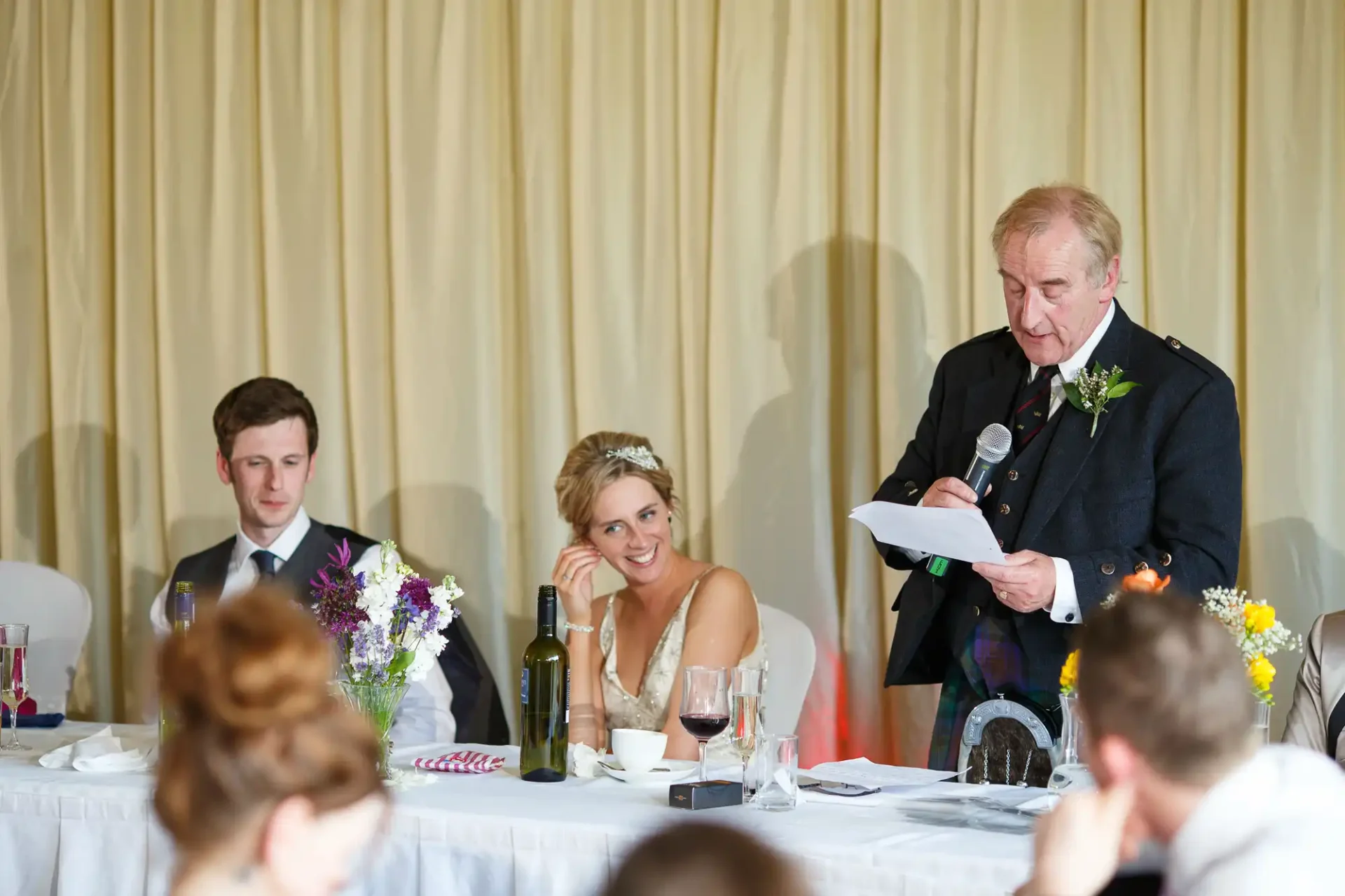 Man giving a speech at a wedding reception, standing next to a seated bride and groom at the head table, with guests listening.