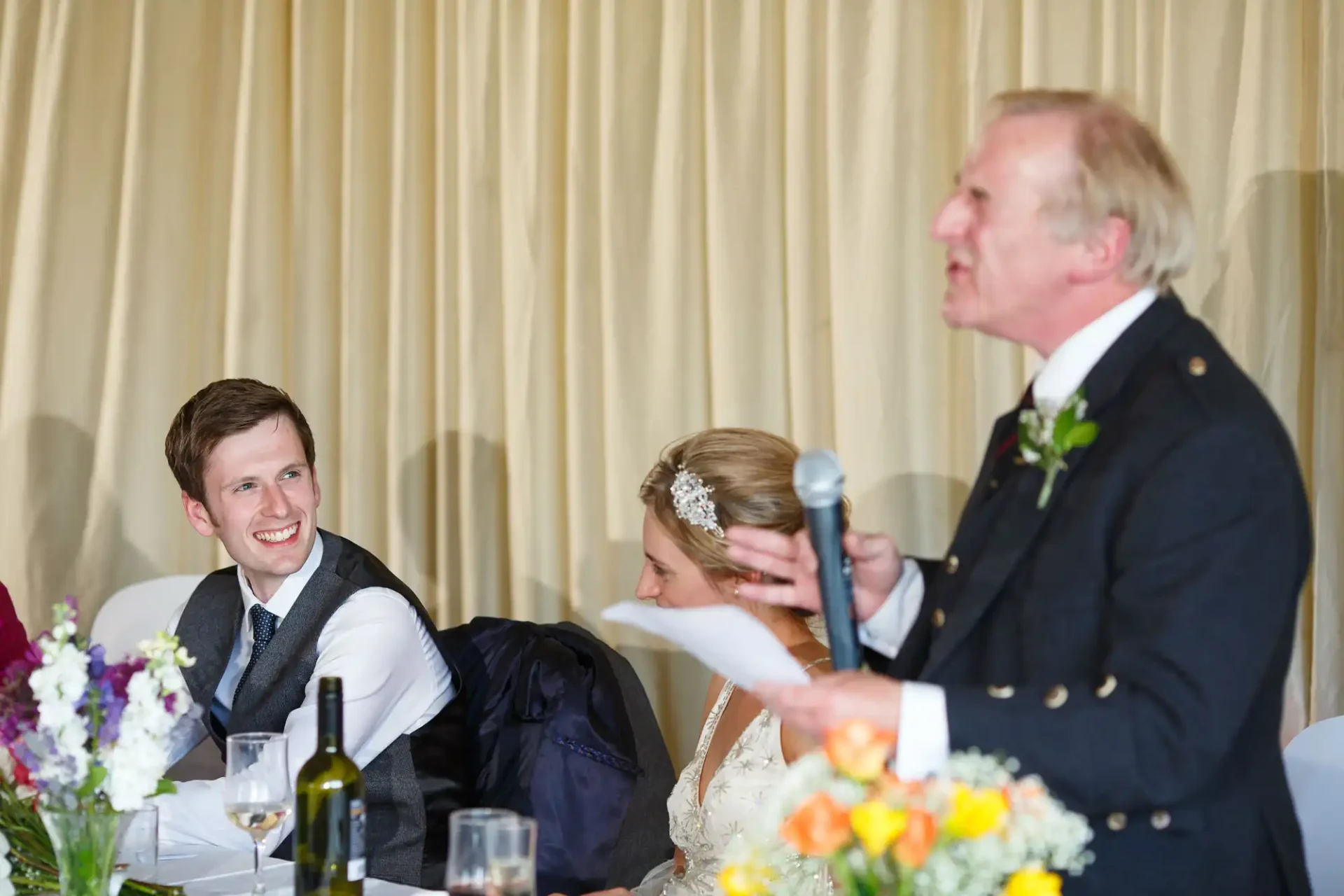 An older man gives a speech at a wedding reception, holding papers, as a smiling young man in a suit and a woman in a white dress look on at a decorated table.