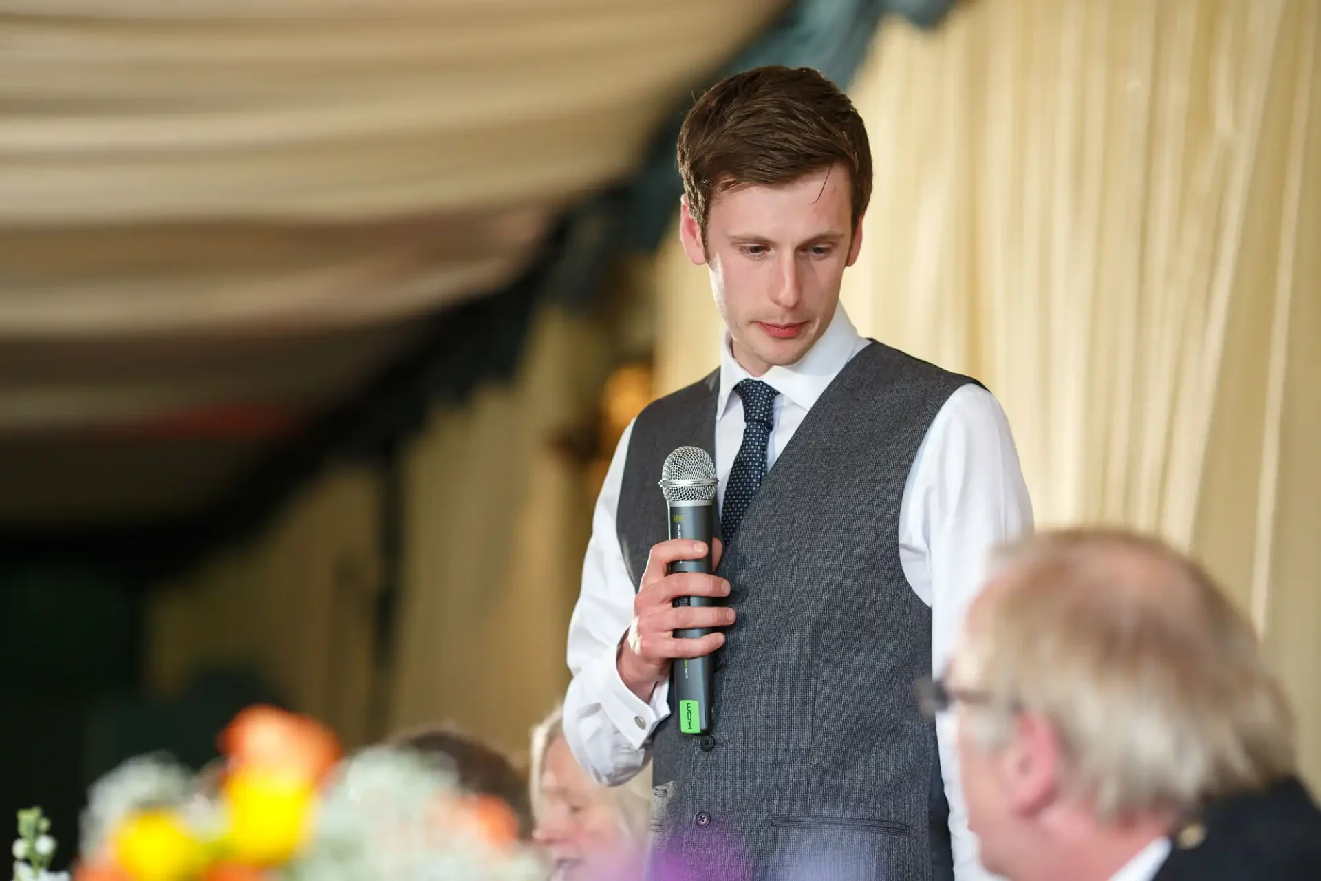 A young man in a vest holding a microphone, giving a speech at an indoor event, with blurred audience members in the foreground.