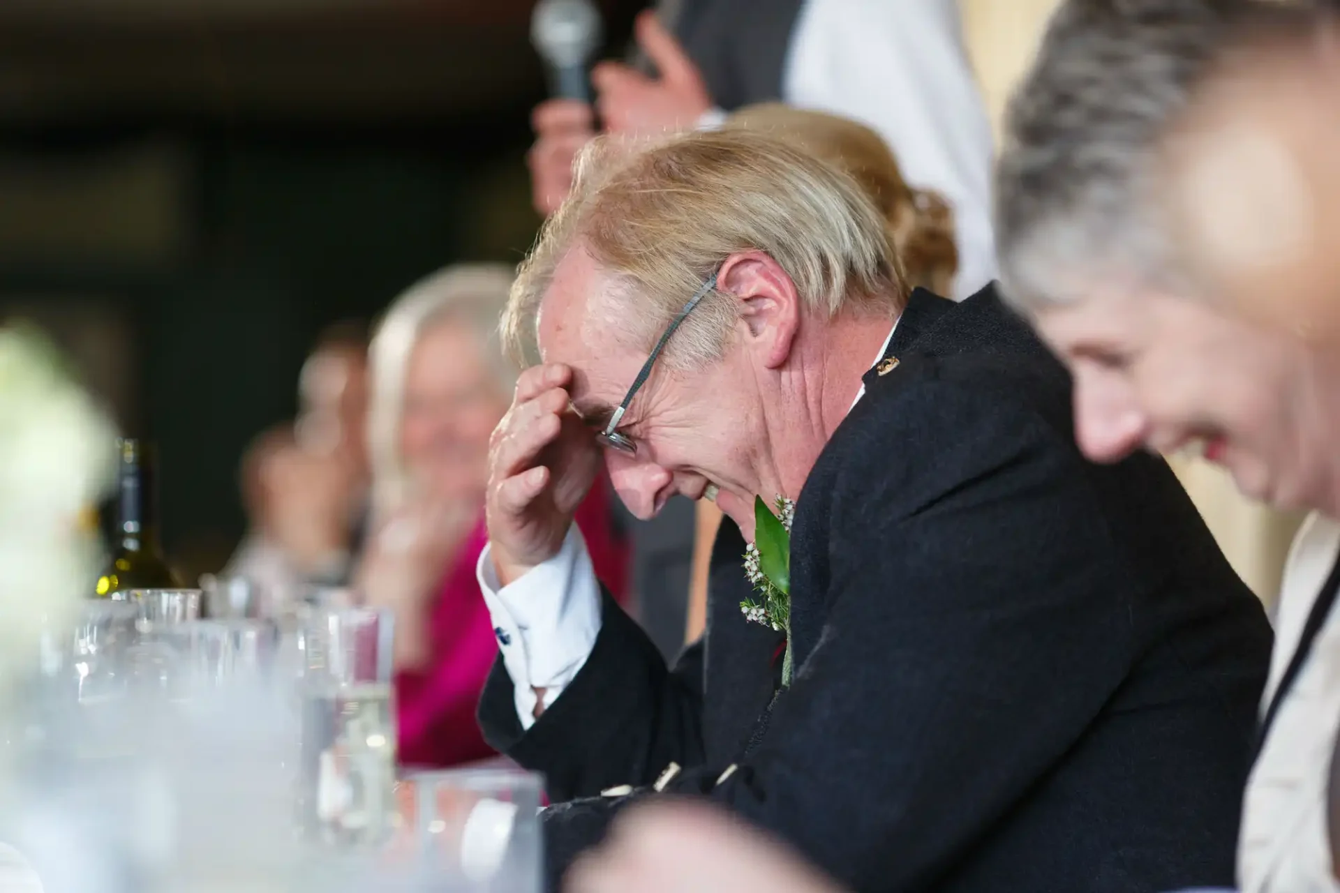A man laughing heartily, holding his glasses, at a wedding reception table with other joyful guests around him.