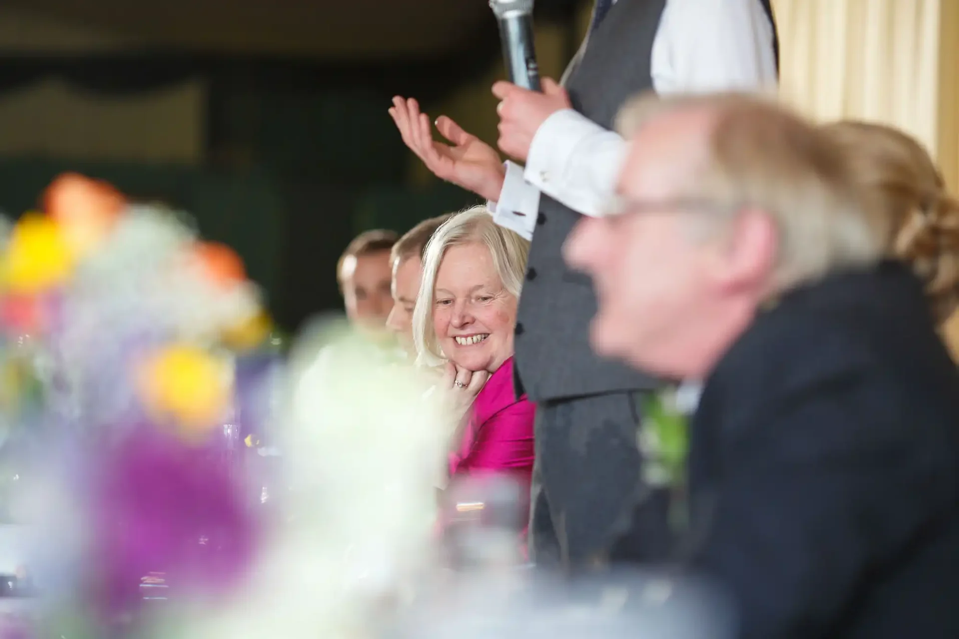 A woman smiles at a wedding reception, looking towards a man holding a microphone, with blurred floral centerpieces in the foreground.