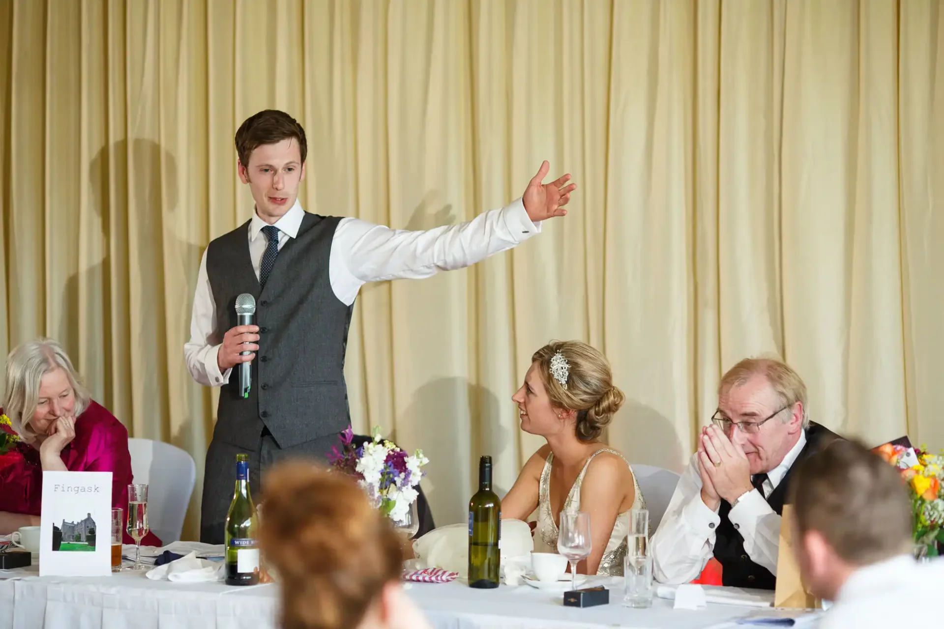 A man giving a speech at a wedding reception, gesturing with his hand, while holding a microphone. the bride and guests are seated at the table, listening.