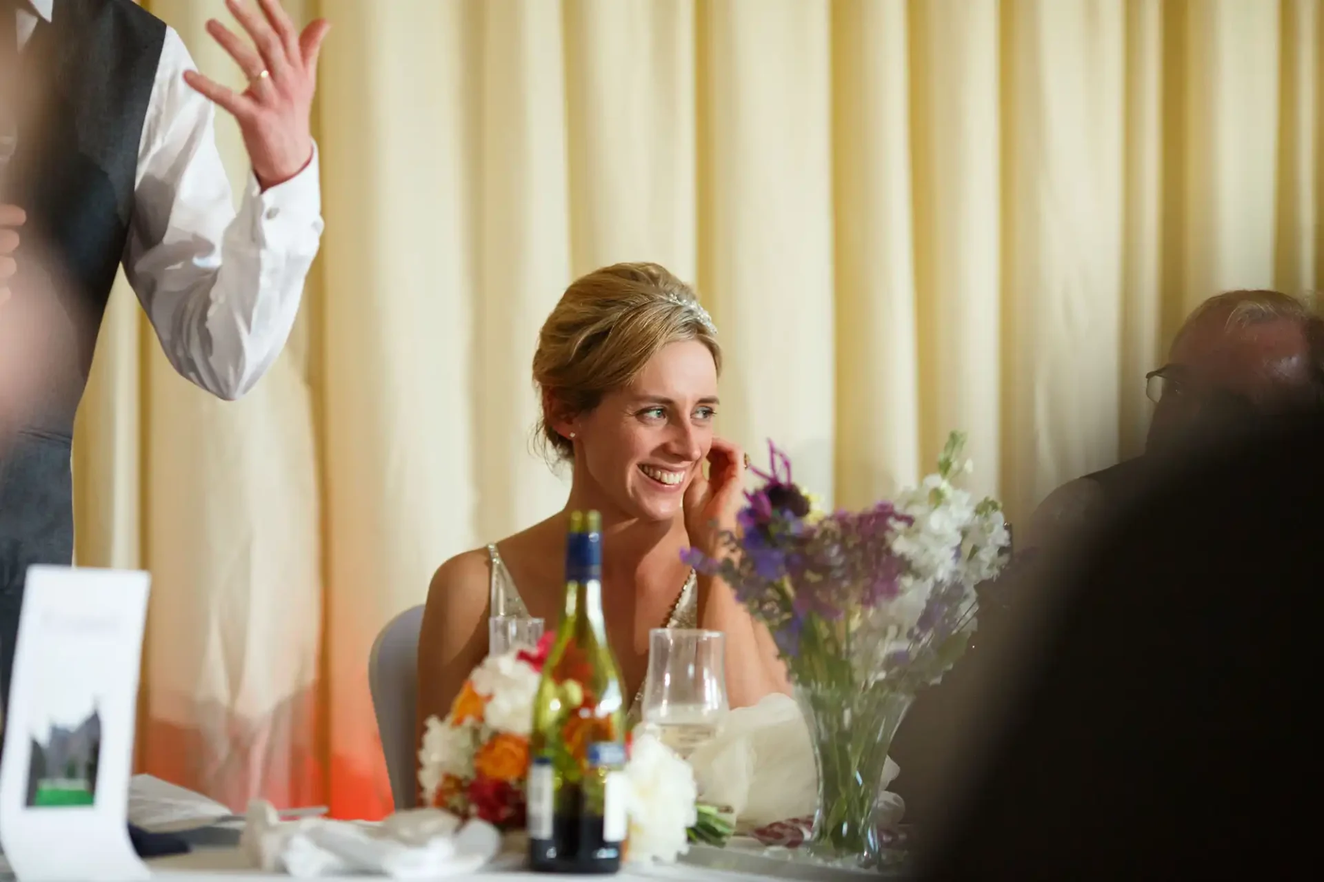 A bride at a wedding reception table, smiling and looking to her left, with guests and decorations around her.