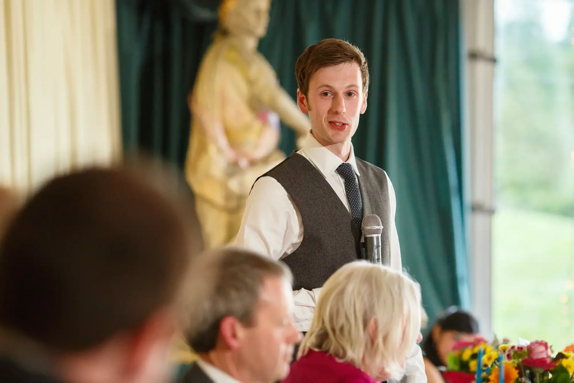 A young man in a vest and tie speaks into a microphone at an event, with guests in the foreground and a statue in the background.
