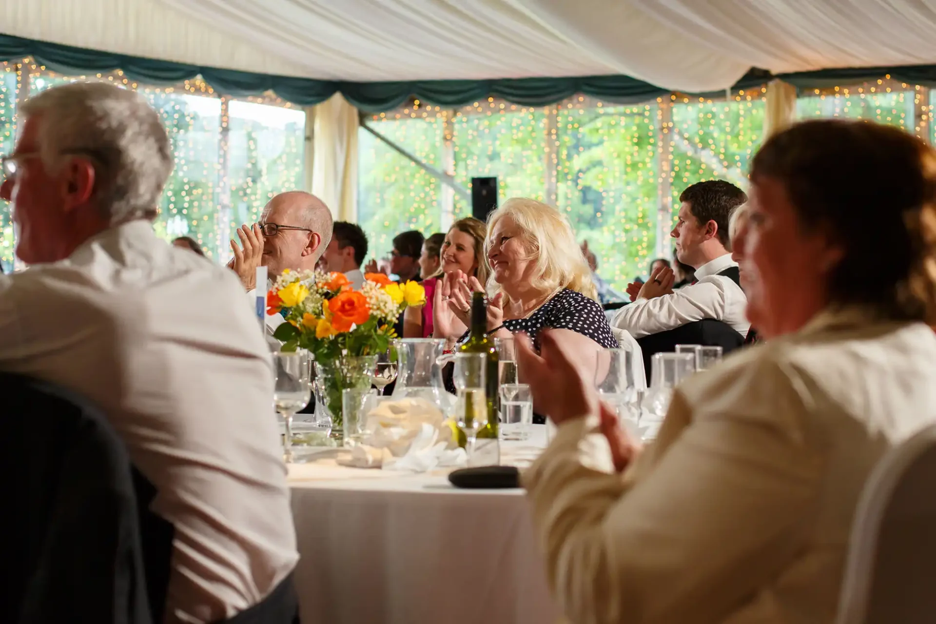 Guests seated at tables in an elegant tent adorned with fairy lights, applauding during a festive event.