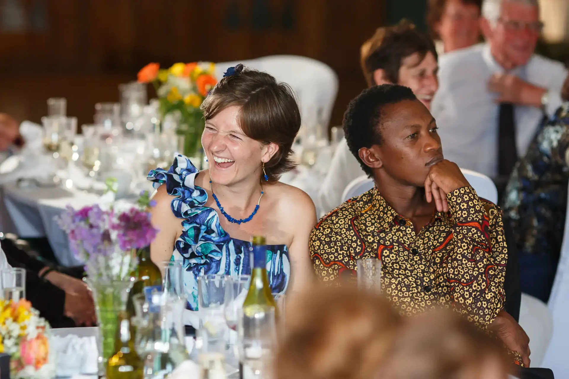A woman laughing joyfully in a blue dress and a man in a patterned shirt attentively watching a speech at a festive event with guests and floral decorations in the background.