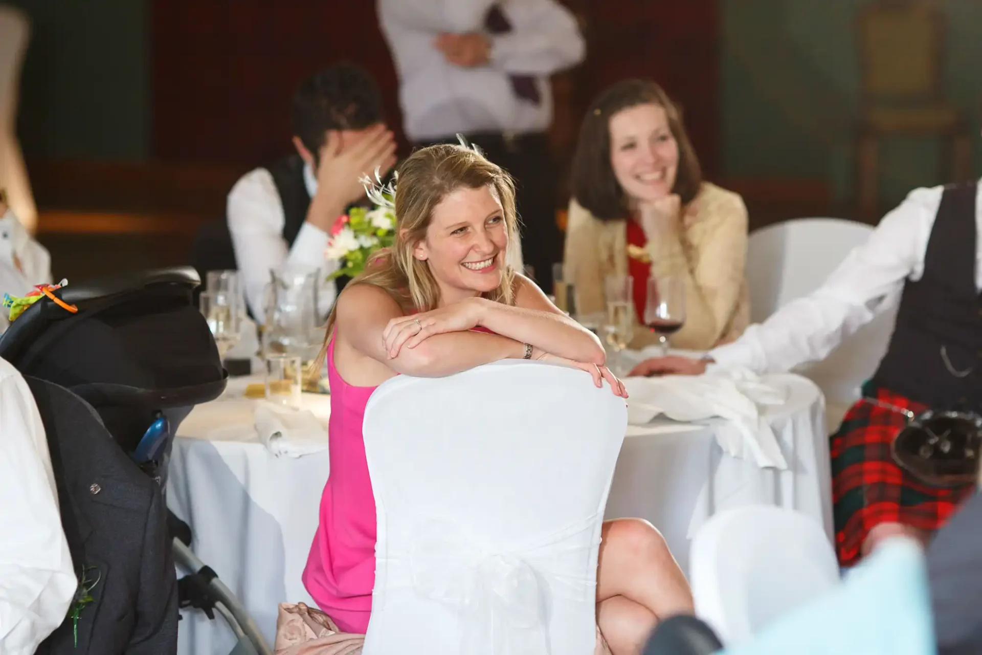 A woman in a white dress smiling at a wedding reception table, with guests in the background, one covering his face and another laughing.