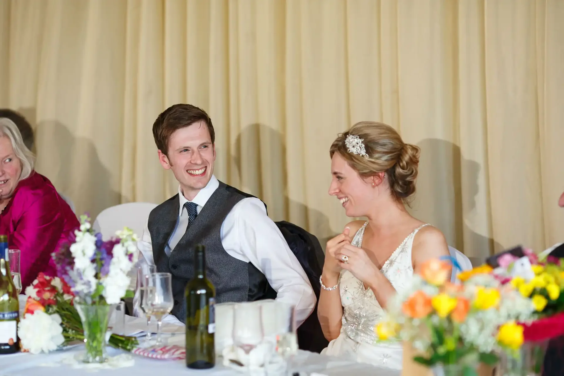 A bride and groom smiling and talking at a wedding reception table decorated with colorful flowers.