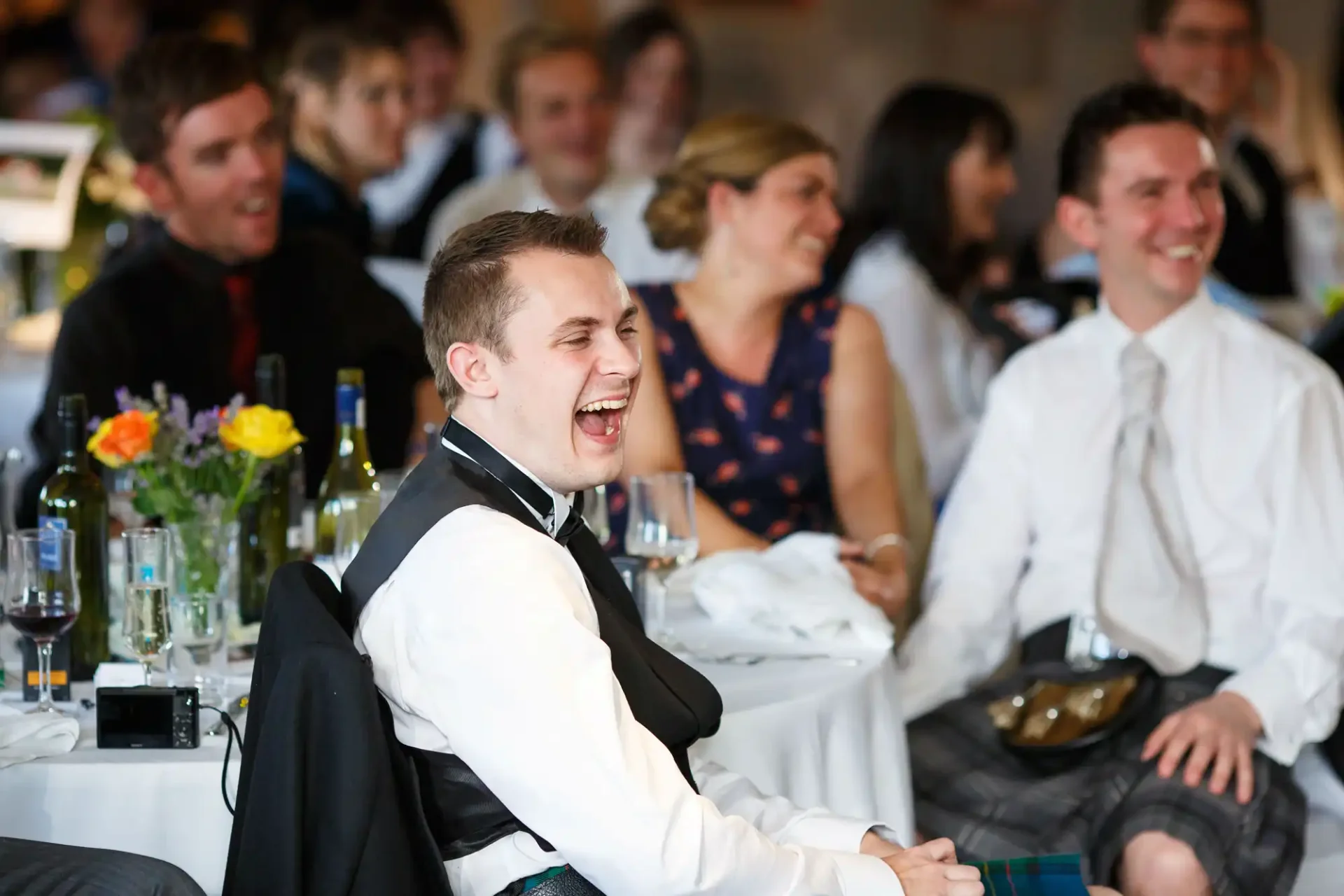 A man in a wheelchair laughs joyously, surrounded by other guests enjoying a festive event, all seated at tables adorned with colorful flowers.