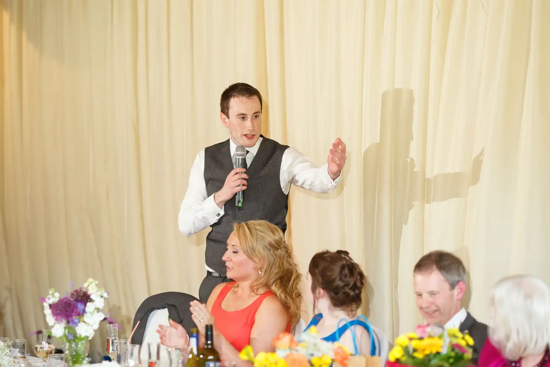 A young man stands giving a speech at a wedding, gesturing with one hand, while holding a microphone with the other. around him, guests seated at tables listen attentively.