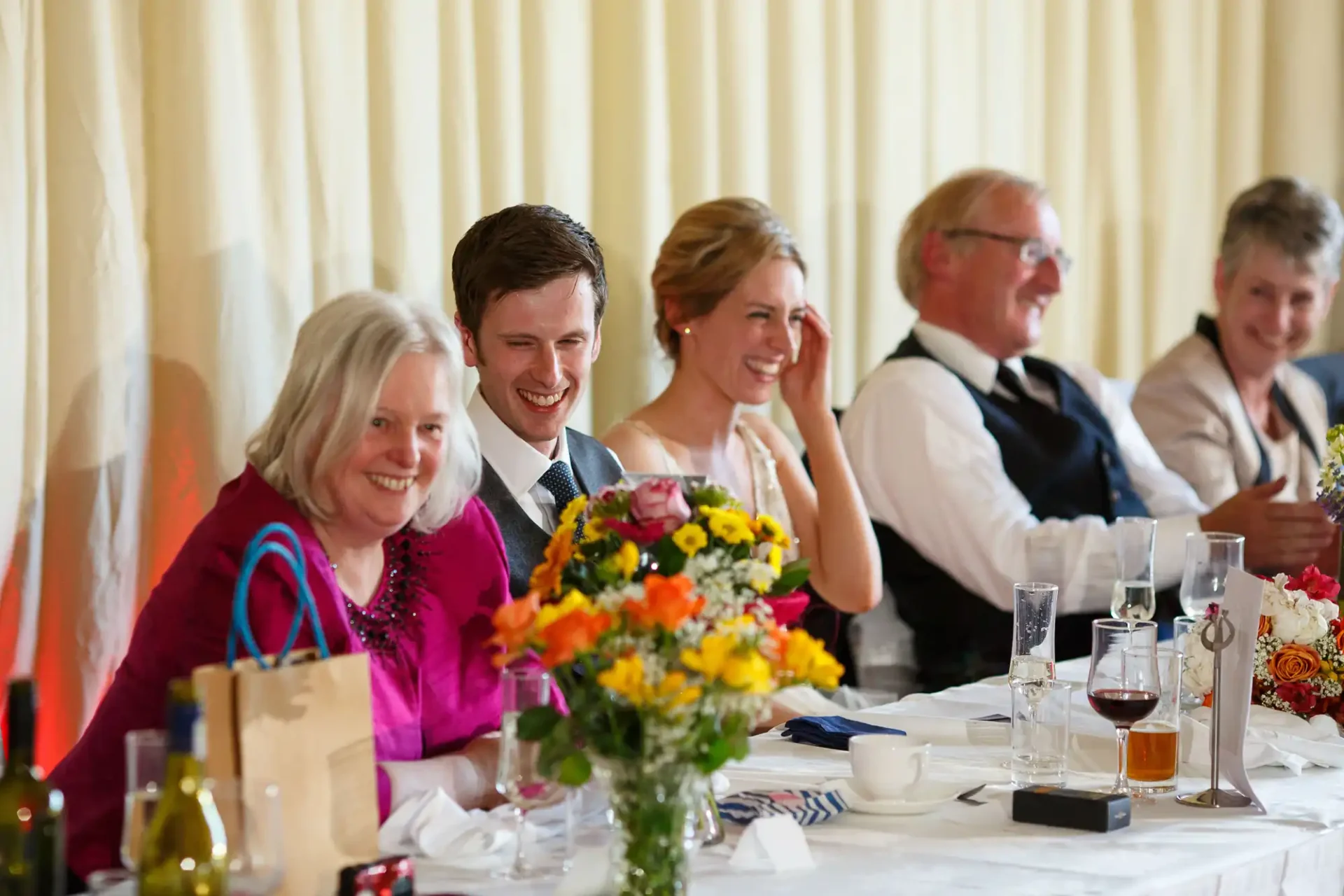 A group of five adults laughing at a table decorated with flowers during a formal event, with beverages and gift bags visible.
