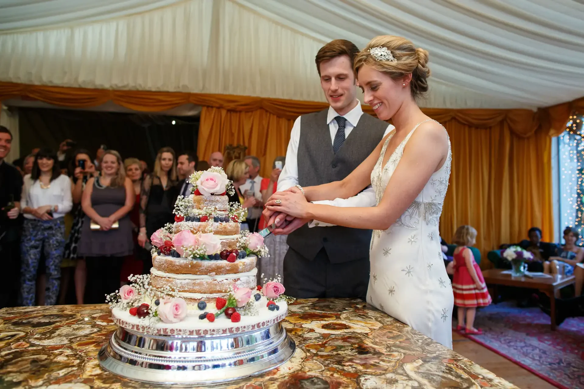 A newlywed couple cuts a multi-tiered wedding cake adorned with pink roses, surrounded by guests in a festively decorated tent.