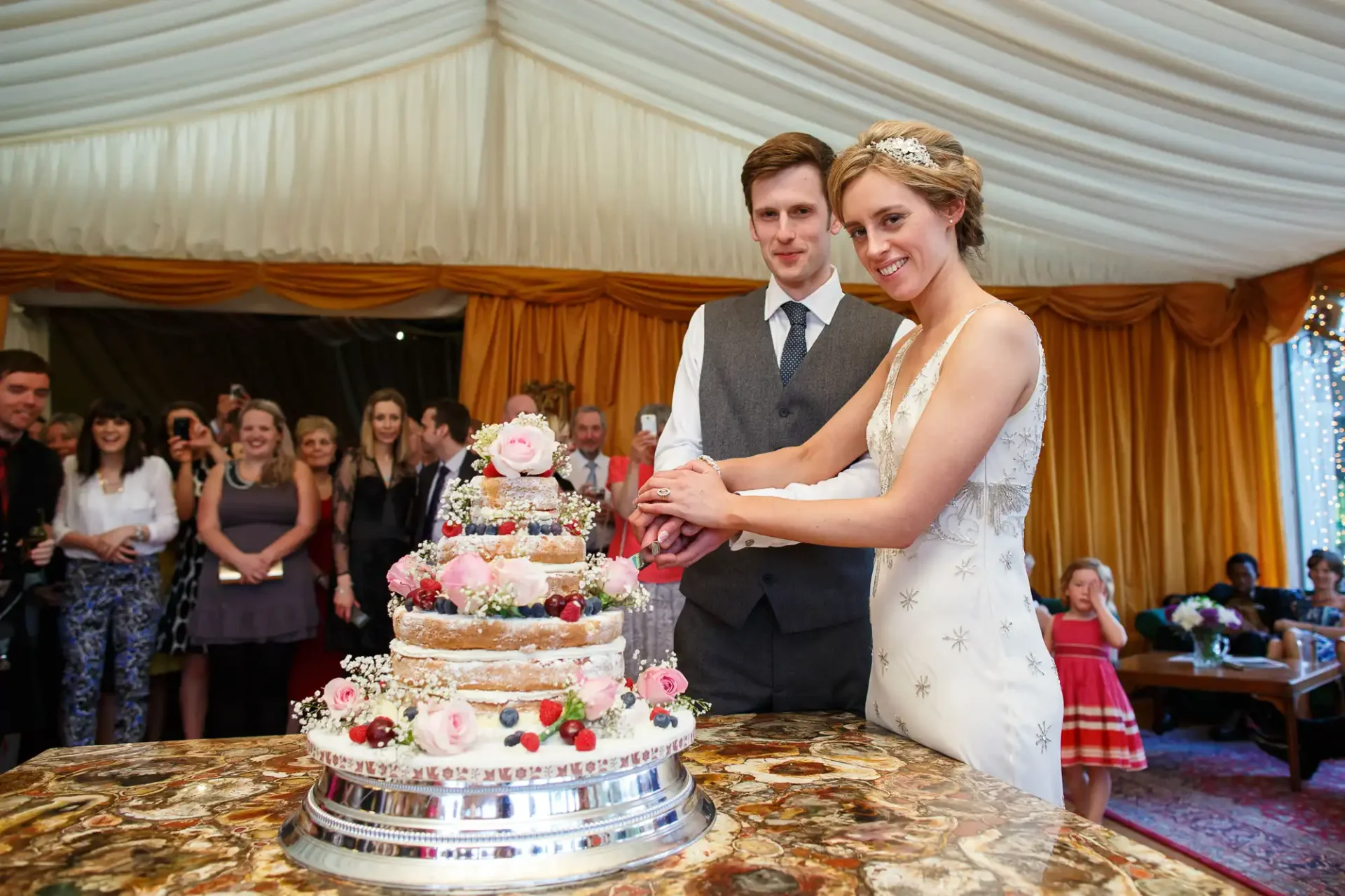 A bride and groom smiling as they cut a multi-tiered wedding cake decorated with flowers, with guests watching in the background.
