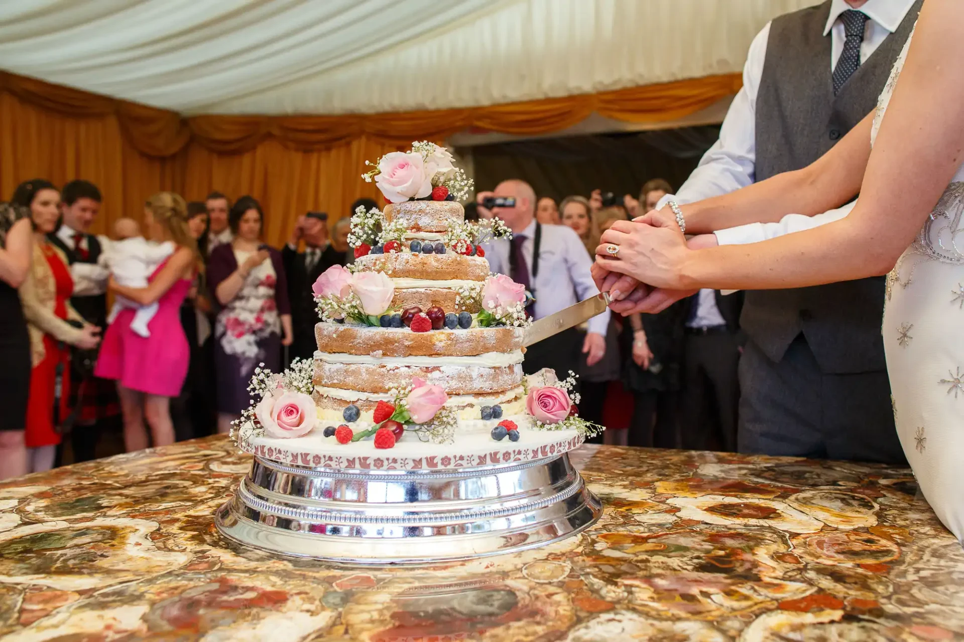 A bride and groom cutting a multi-tiered wedding cake adorned with fresh flowers, surrounded by guests in a tent.
