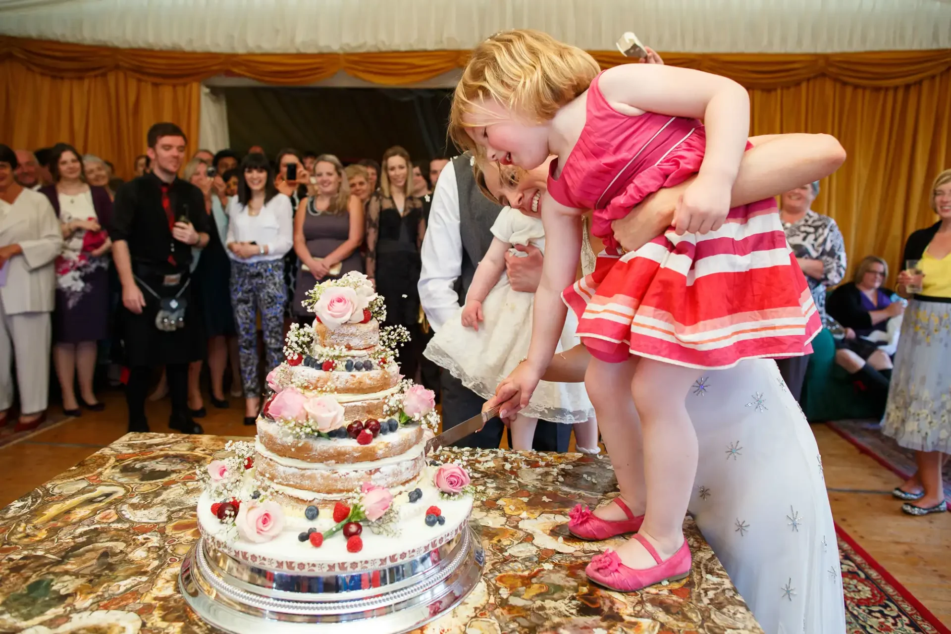 A young girl in a striped dress excitedly reaches for a tiered wedding cake at a reception, held by a smiling woman, with guests watching.