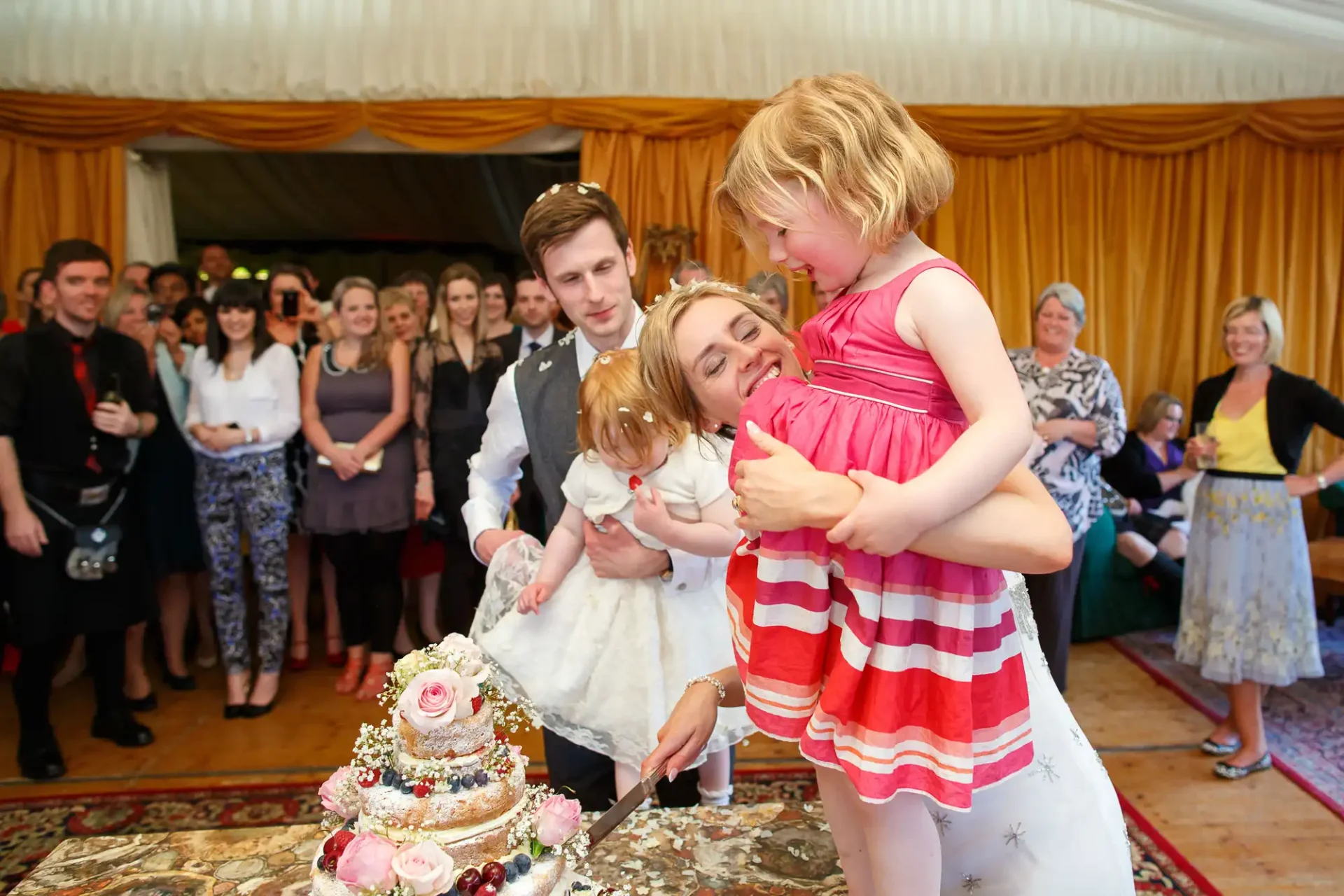 A mother with a toy doll kissing a young girl in a striped dress at a wedding, with guests observing and a man beside them looking on.