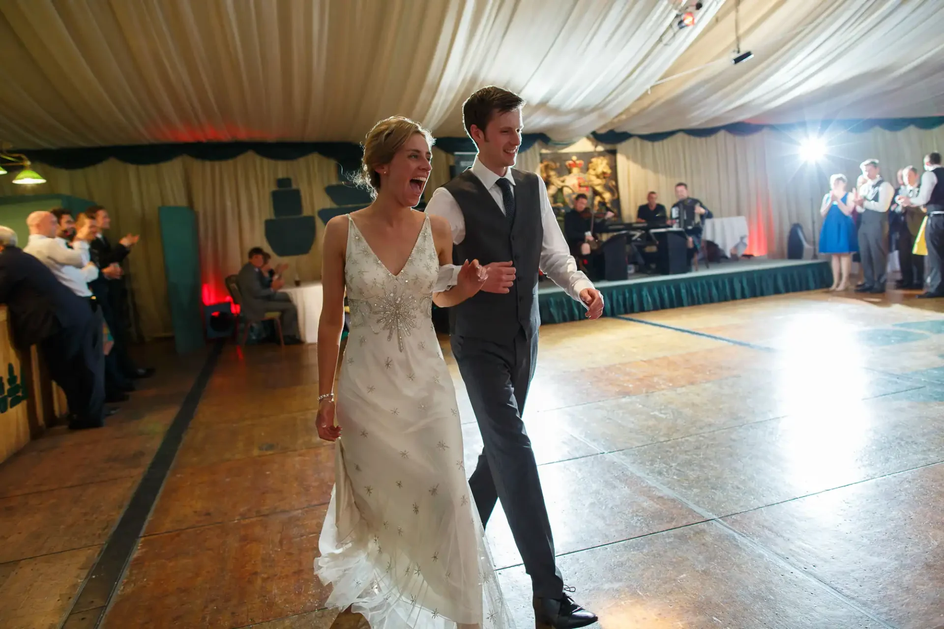 A bride and groom walk hand in hand, smiling, across a dance floor with guests watching in the background at a wedding reception.