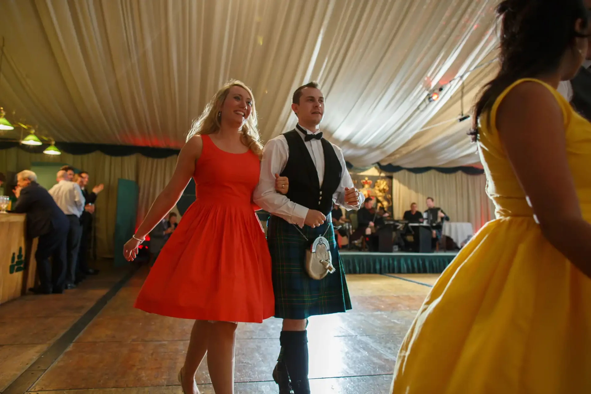 A man in a kilt and a woman in a red dress walking hand in hand at a festive event under a large tent.