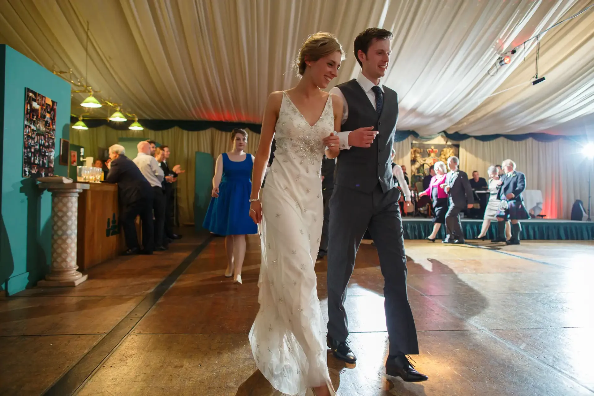 A young couple dances happily at a wedding reception in a tented venue, with guests in the background.
