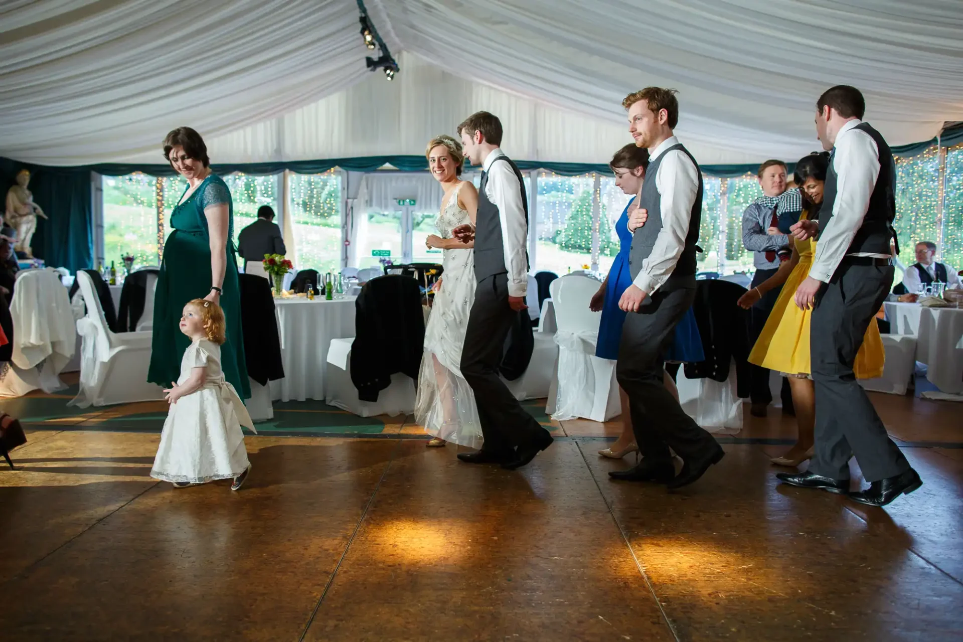 Guests dancing at a wedding reception, including a young girl looking up at a smiling woman in a long dress.