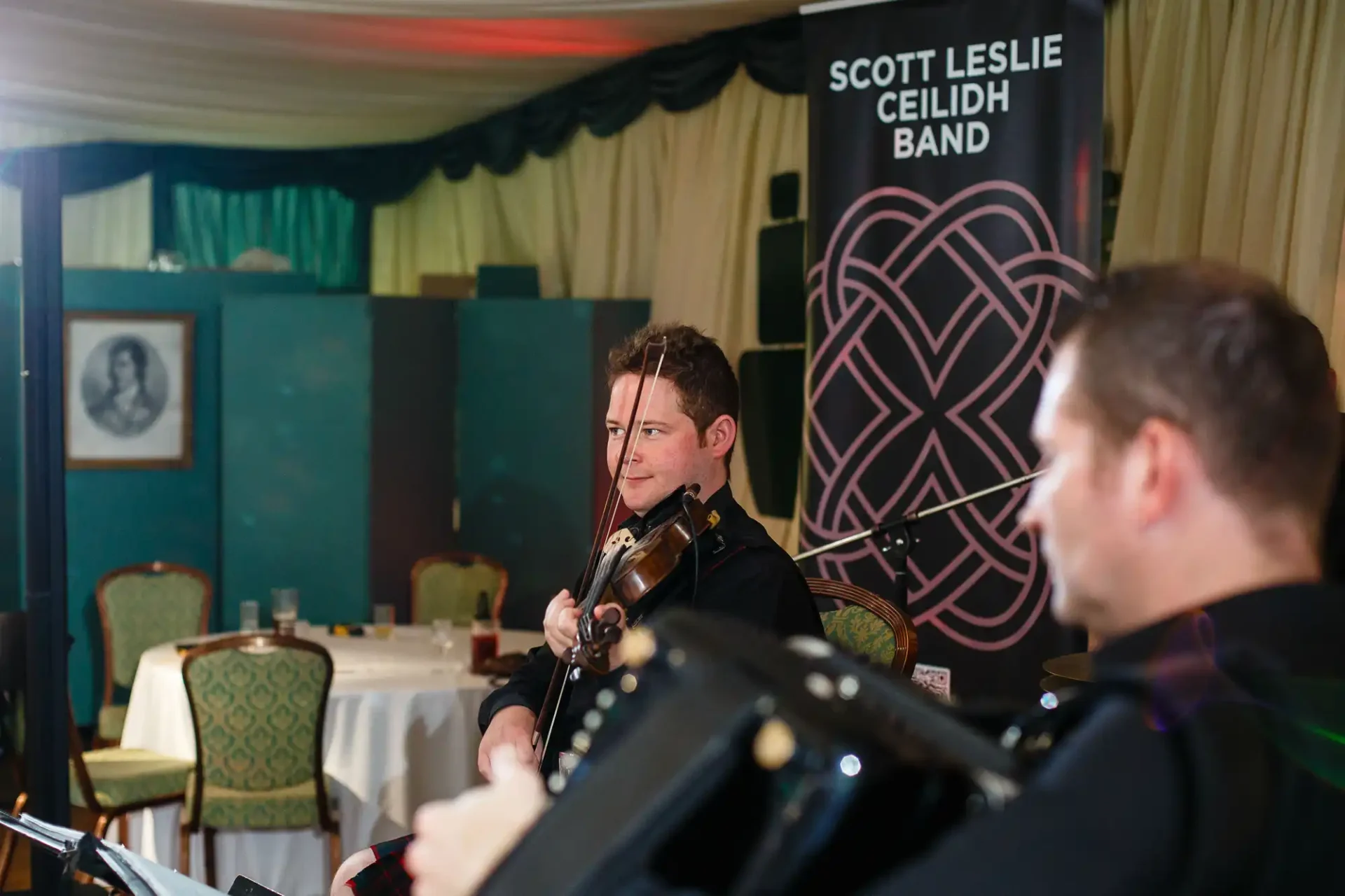 Violinist performing on stage with the scott leslie ceilidh band, viewed over the shoulder of another band member in a warmly lit room with event decor.