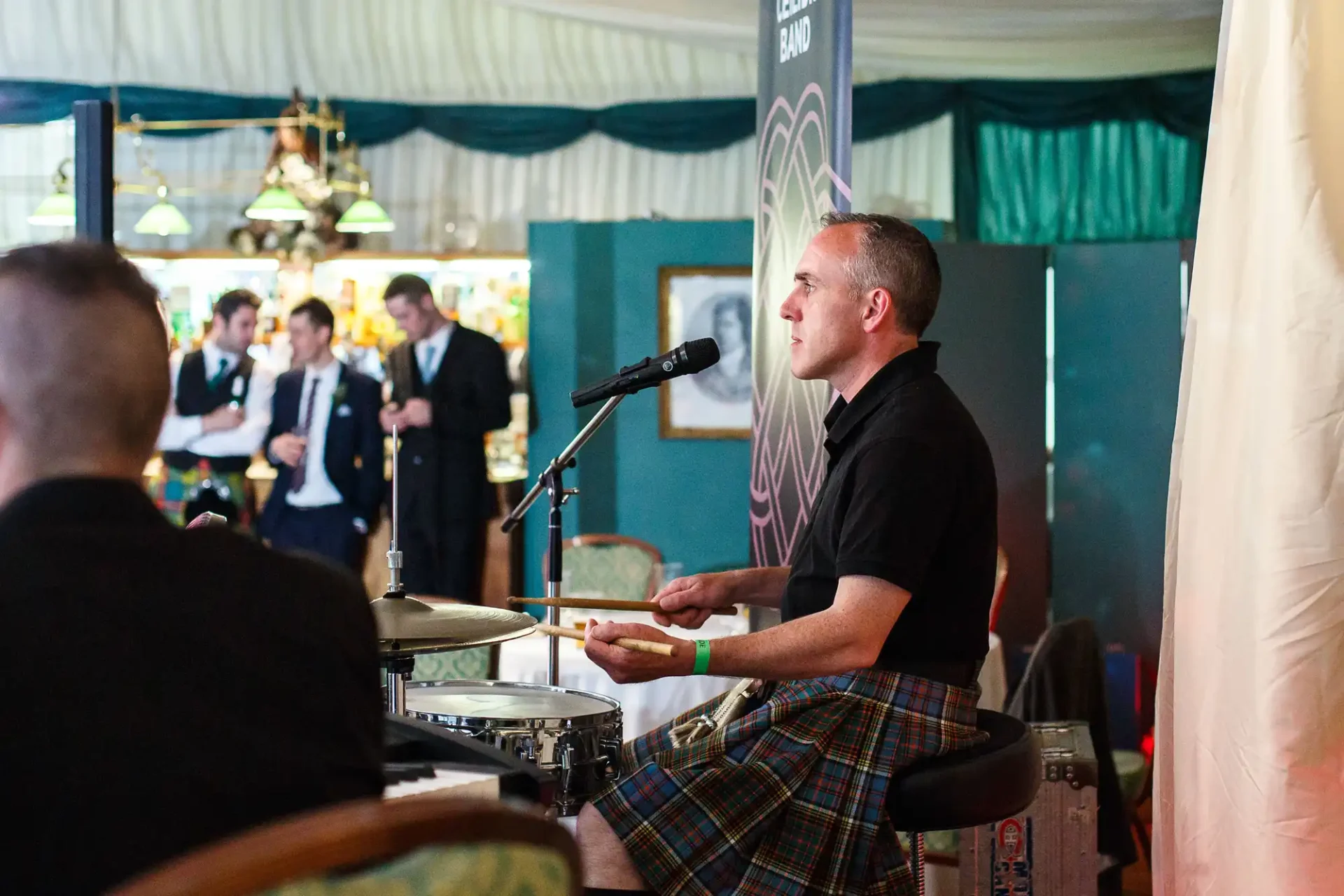 A drummer in a kilt performs at an indoor event, focused intently on his drum set, with audience members and musicians in the background.