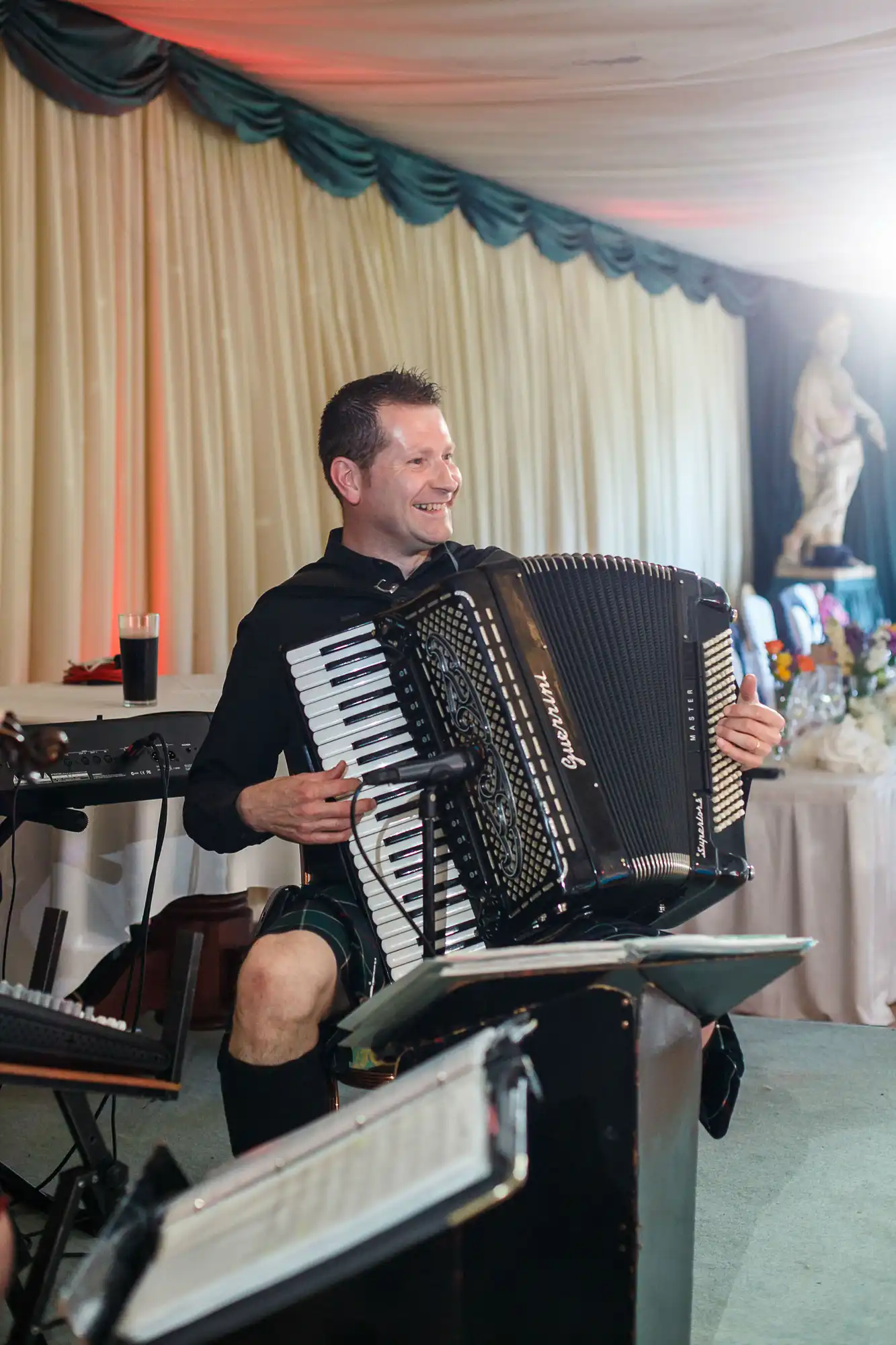 A man smiling while playing an accordion at an indoor event, with a keyboard and decorated tables in the background.
